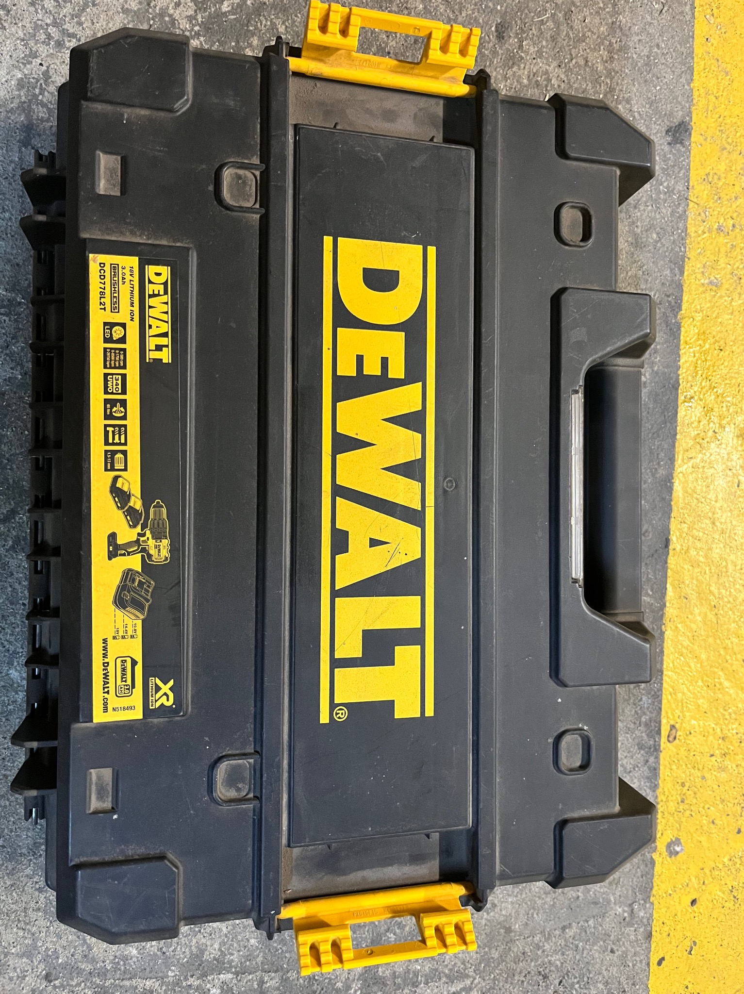 Czarna skrzynka narzędziowa DeWalt z żółtymi akcentami, leżąca na szarej powierzchni z żółtą linią. Widoczne logo i specyfikacje narzędzia.