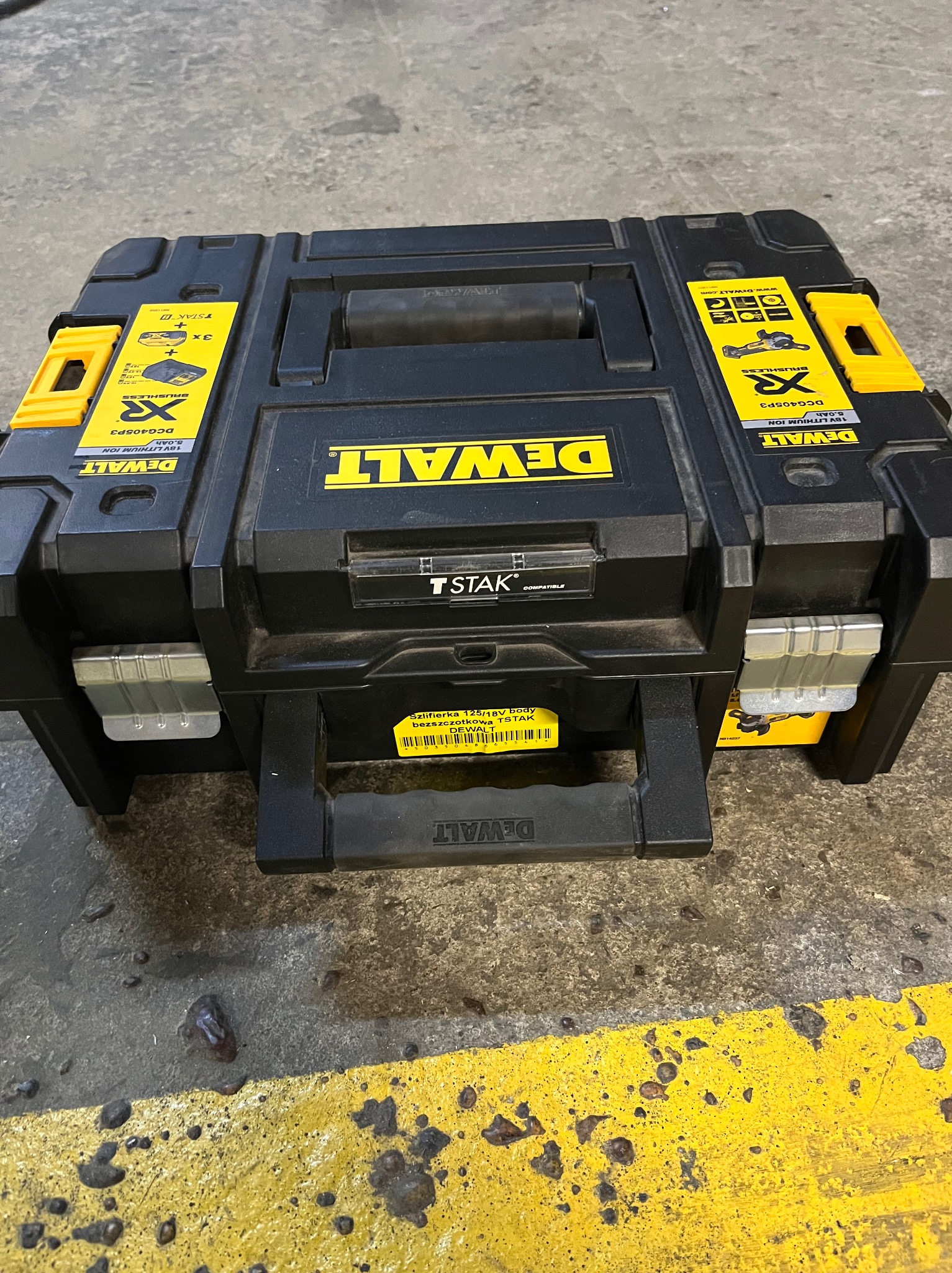 Czarna skrzynka narzędziowa DeWalt TSTAK na podłodze z żółtą linią. Widoczne logo DeWalt i oznaczenia modelu szlifierki bezszczotkowej 125/18V.