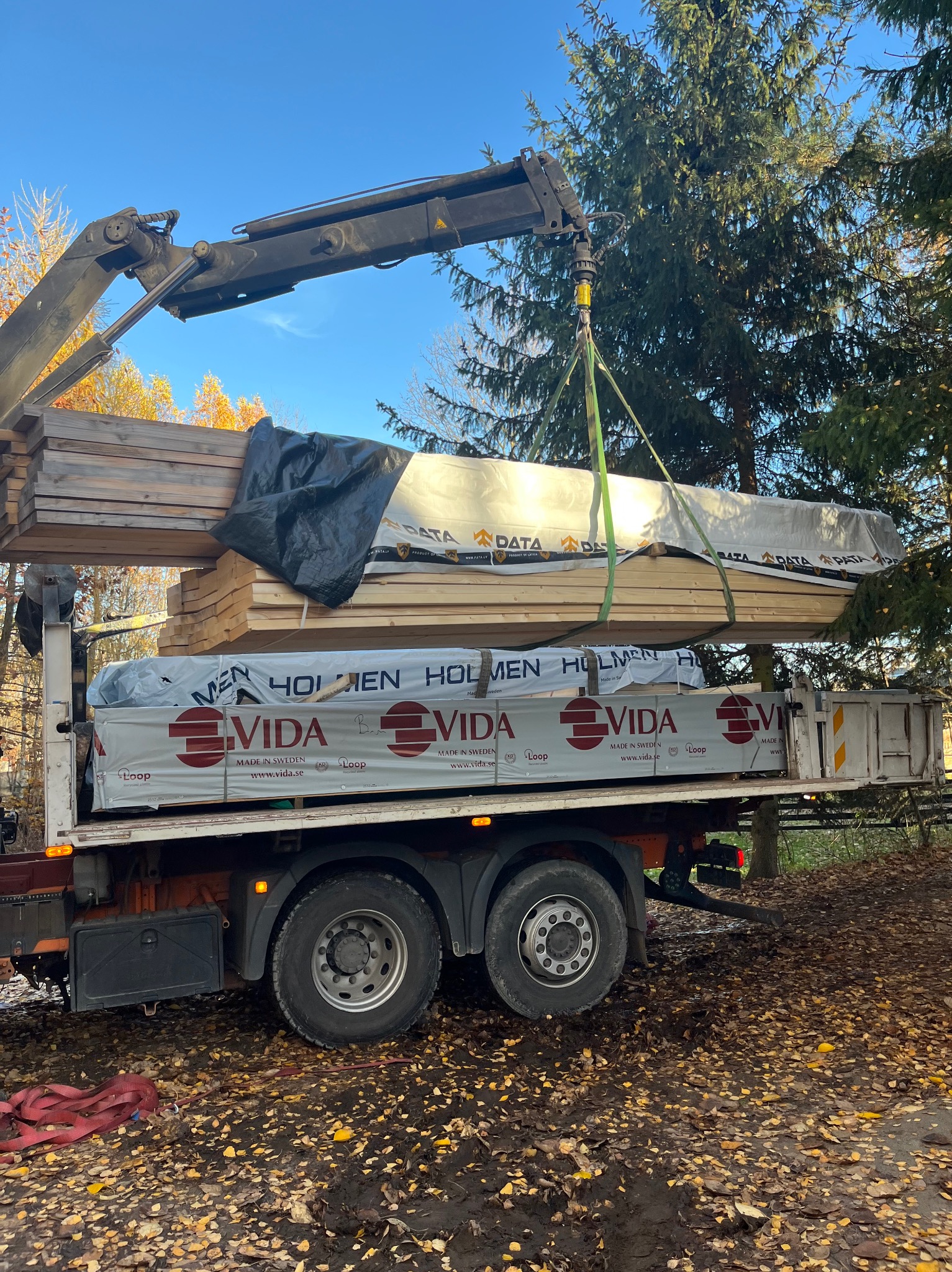 Transport drewna VIDA na budowę domu szkieletowego. Żuraw podnosi pakiet desek z ciężarówki na tle drzew i błękitnego nieba.