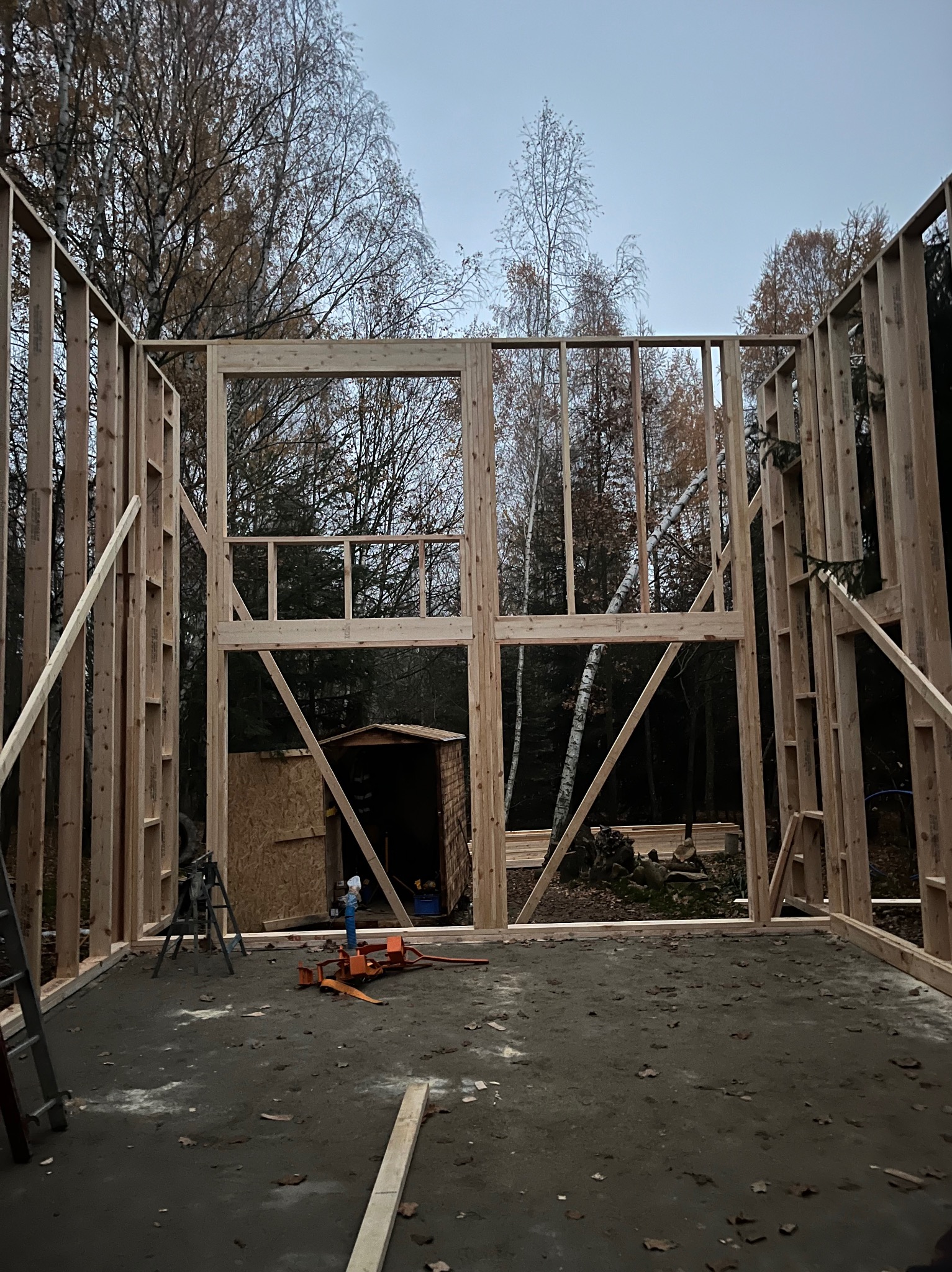 Wnętrze w trakcie budowy domu szkieletowego. Widoczne drewniane ramy ścian, mała budka w tle i narzędzia na betonowej podłodze. Surowy stan, jesienna sceneria za oknami.