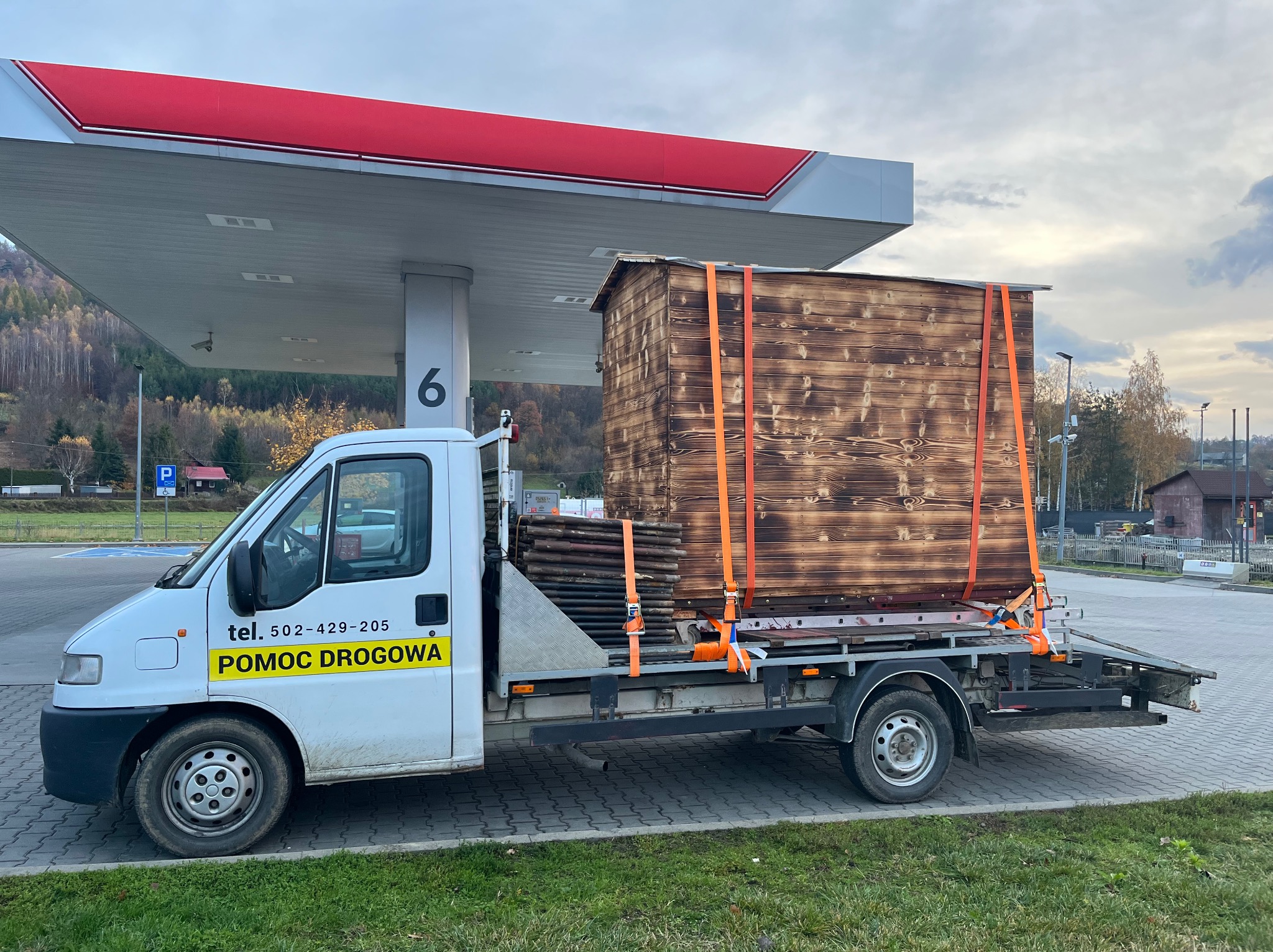 Transport modułu domu szkieletowego na lawecie pomocy drogowej, widok z boku na tle stacji benzynowej. Moduł zabezpieczony pomarańczowymi pasami.