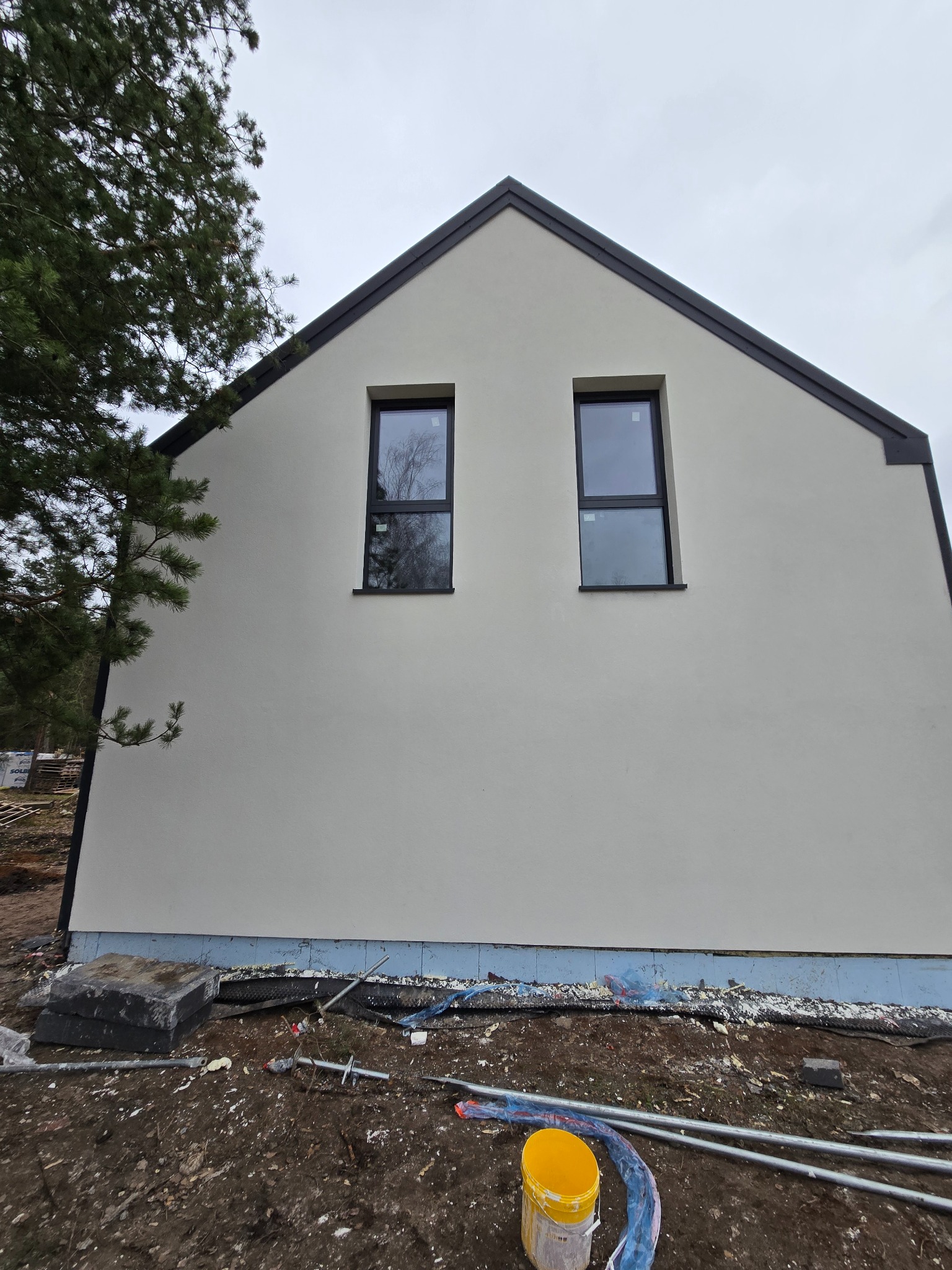 Świeżo otynkowana ściana domu z dwoma oknami, widoczne materiały budowlane i drzewo z boku. Elewacja w jasnym odcieniu, czarne obramowania okien i dachu.