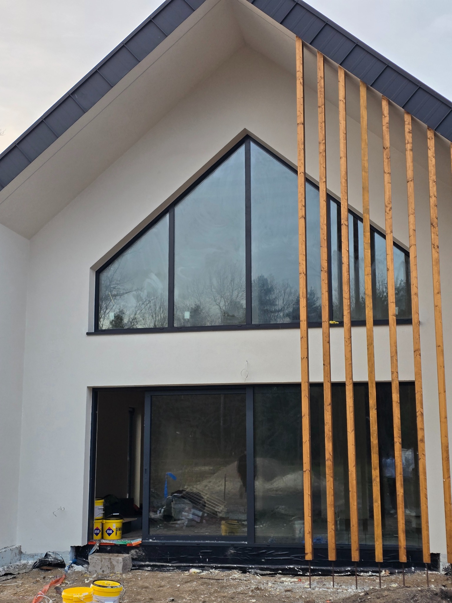 Nowoczesny dom z białymi ścianami, dużymi oknami i drewnianymi elementami dekoracyjnymi. Widoczne detale elewacji i otoczenie w trakcie prac wykończeniowych.