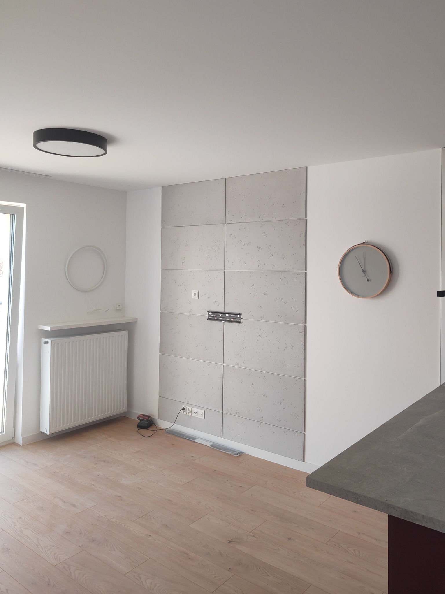 Pokój po wykończeniu: panele ścienne z szarego betonu architektonicznego, drewniana podłoga, białe ściany, grzejnik i okrągły zegar. Widoczne gniazdka i uchwyt na telewizor.