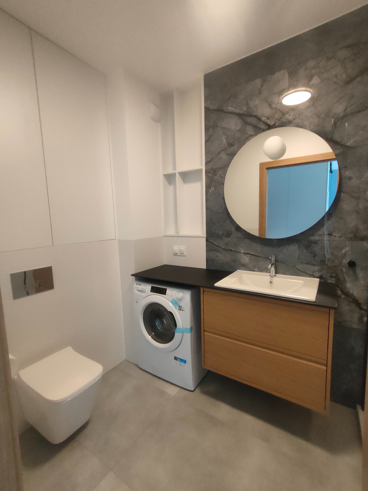 Nowoczesna łazienka z marmurową ścianą, okrągłym lustrem, drewnianą szafką pod umywalką, pralką i podwieszaną toaletą. Minimalistyczny design, szare płytki podłogowe.