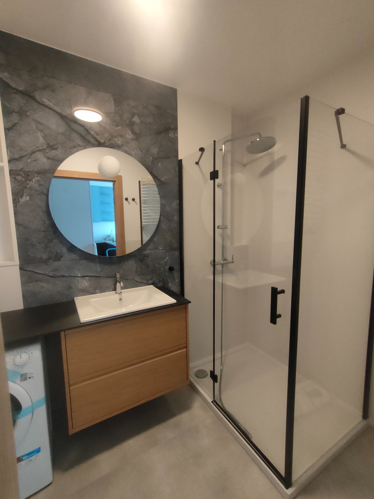 Nowoczesna łazienka z kabiną prysznicową typu walk-in, umywalką na szafce z drewnianym frontem i okrągłym lustrem na tle ściany z efektem marmuru.