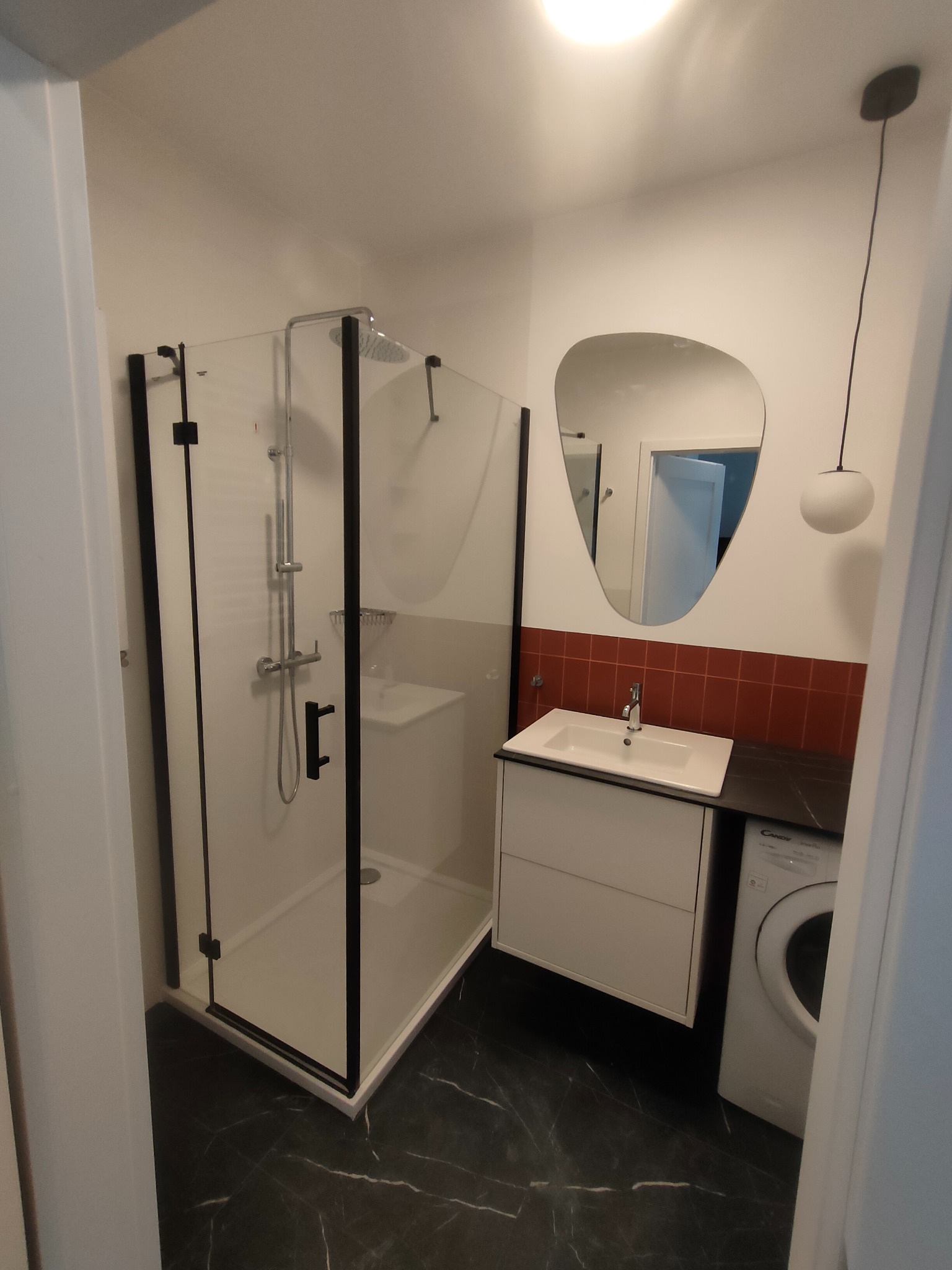 Nowoczesna łazienka po remoncie: kabina prysznicowa z czarną ramą, umywalka z szafką, lustro o nieregularnym kształcie, płytki w odcieniach czerwieni i pralka.