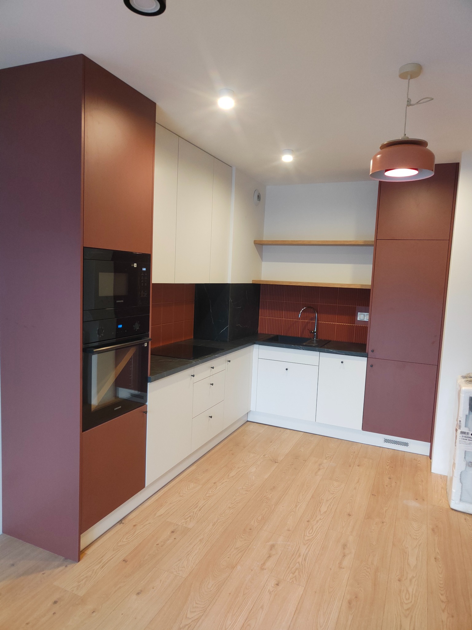 Nowoczesna kuchnia w odcieniach bieli i bordo, z drewnianą podłogą. Widoczne zabudowane sprzęty AGD i oświetlenie punktowe. Uwagę przykuwa nietypowa kolorystyka płytek.