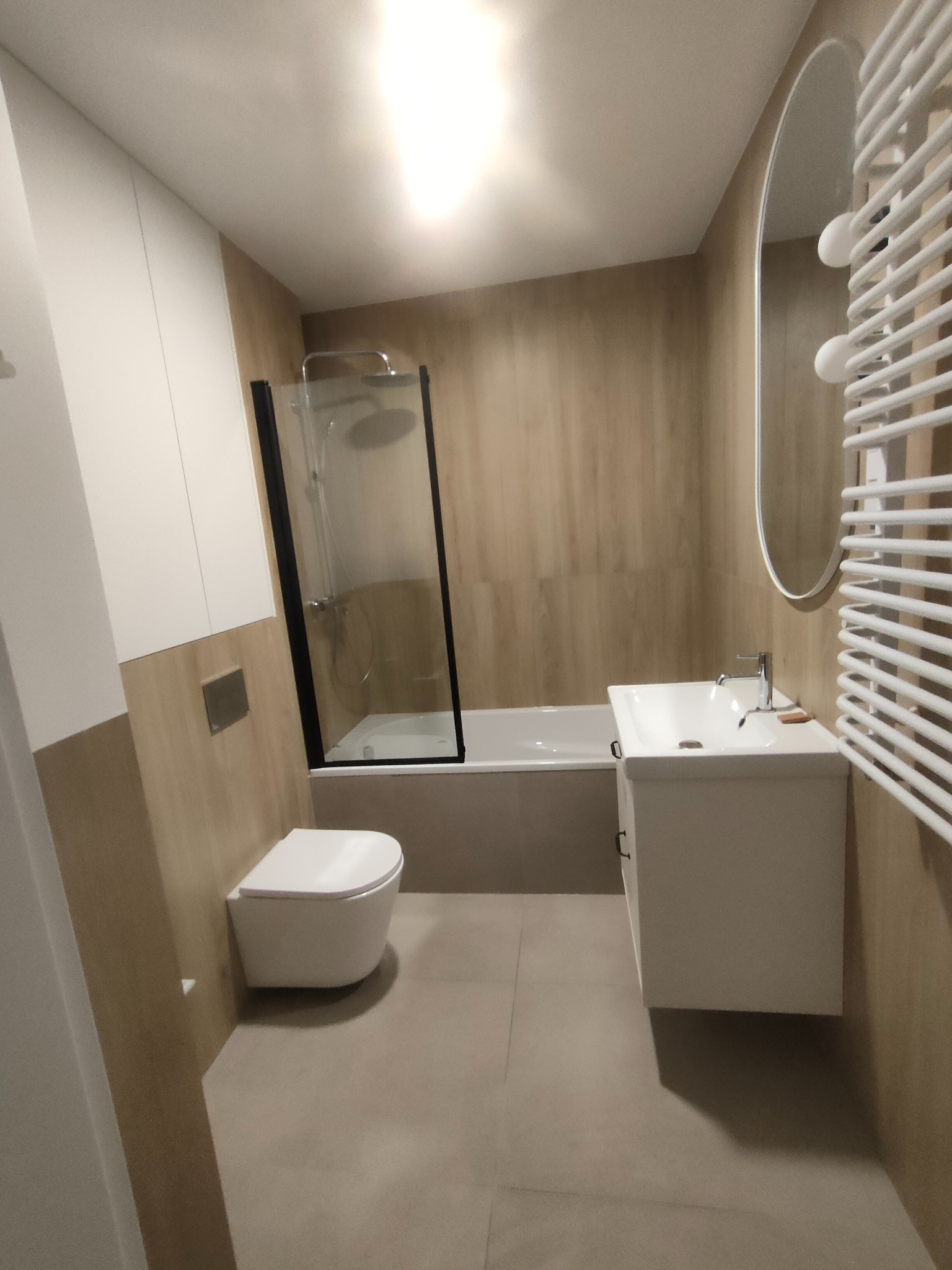 Nowoczesna łazienka z kabiną prysznicową, umywalką z szafką i podwieszaną toaletą. Beżowe płytki imitujące drewno, białe meble i grzejnik drabinkowy.