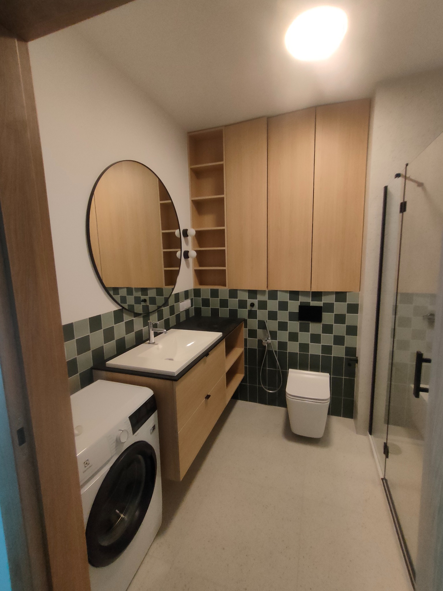 Nowoczesna łazienka z drewnianymi szafkami, okrągłym lustrem i pralką. Uwagę przykuwają zielone płytki w szachownicę nadające wnętrzu unikalny charakter.