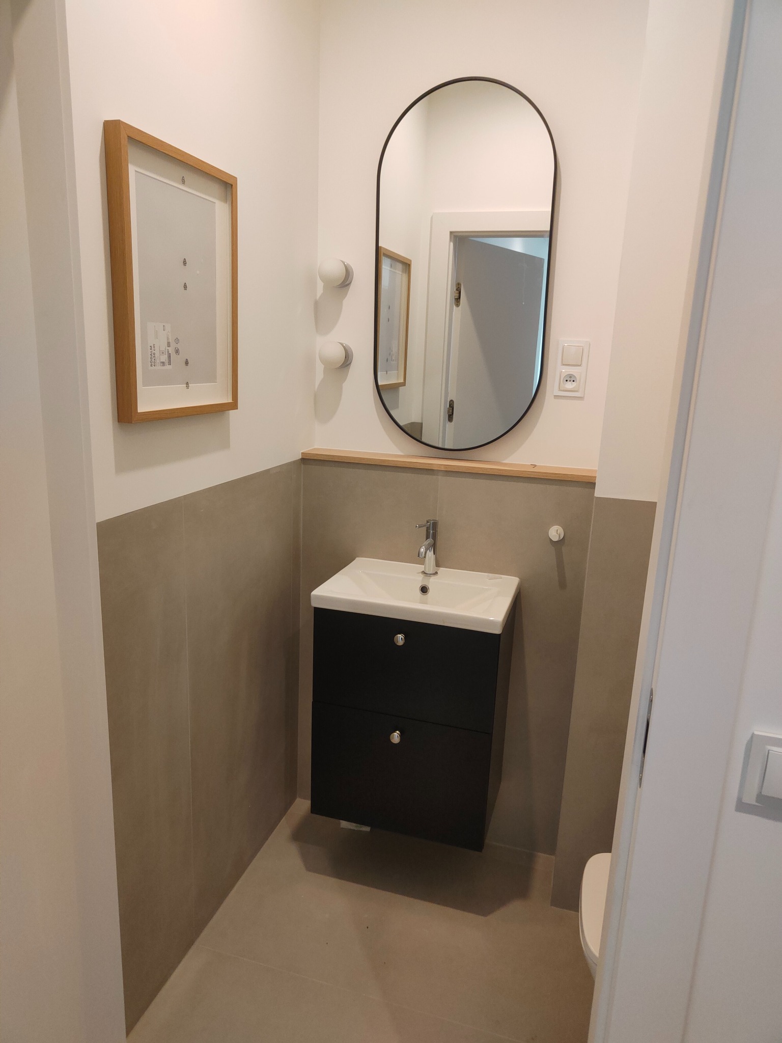 Nowoczesna łazienka z umywalką w szafce, owalnym lustrem i szarą glazurą do połowy ściany. Minimalistyczny design, jasne oświetlenie, rama obrazu na ścianie.