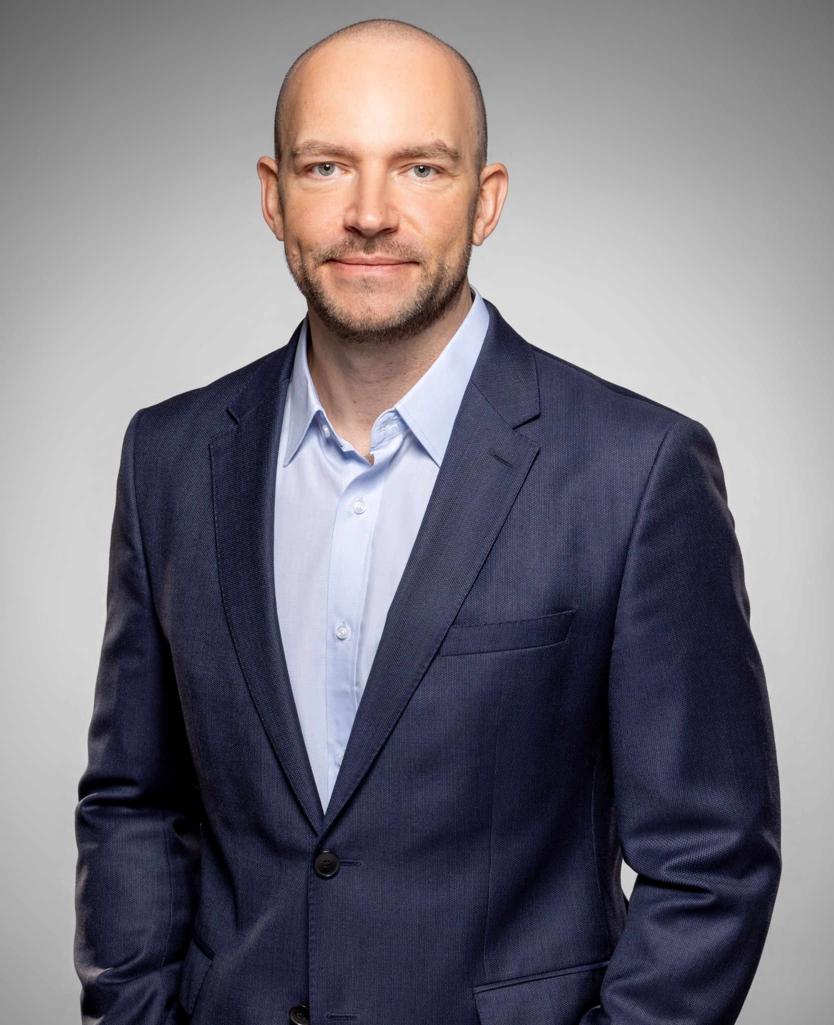 Portret biznesowy: mężczyzna w granatowej marynarce i błękitnej koszuli na jasnoszarym tle. Profesjonalny wygląd, subtelny uśmiech, bezpośrednie spojrzenie w obiektyw.