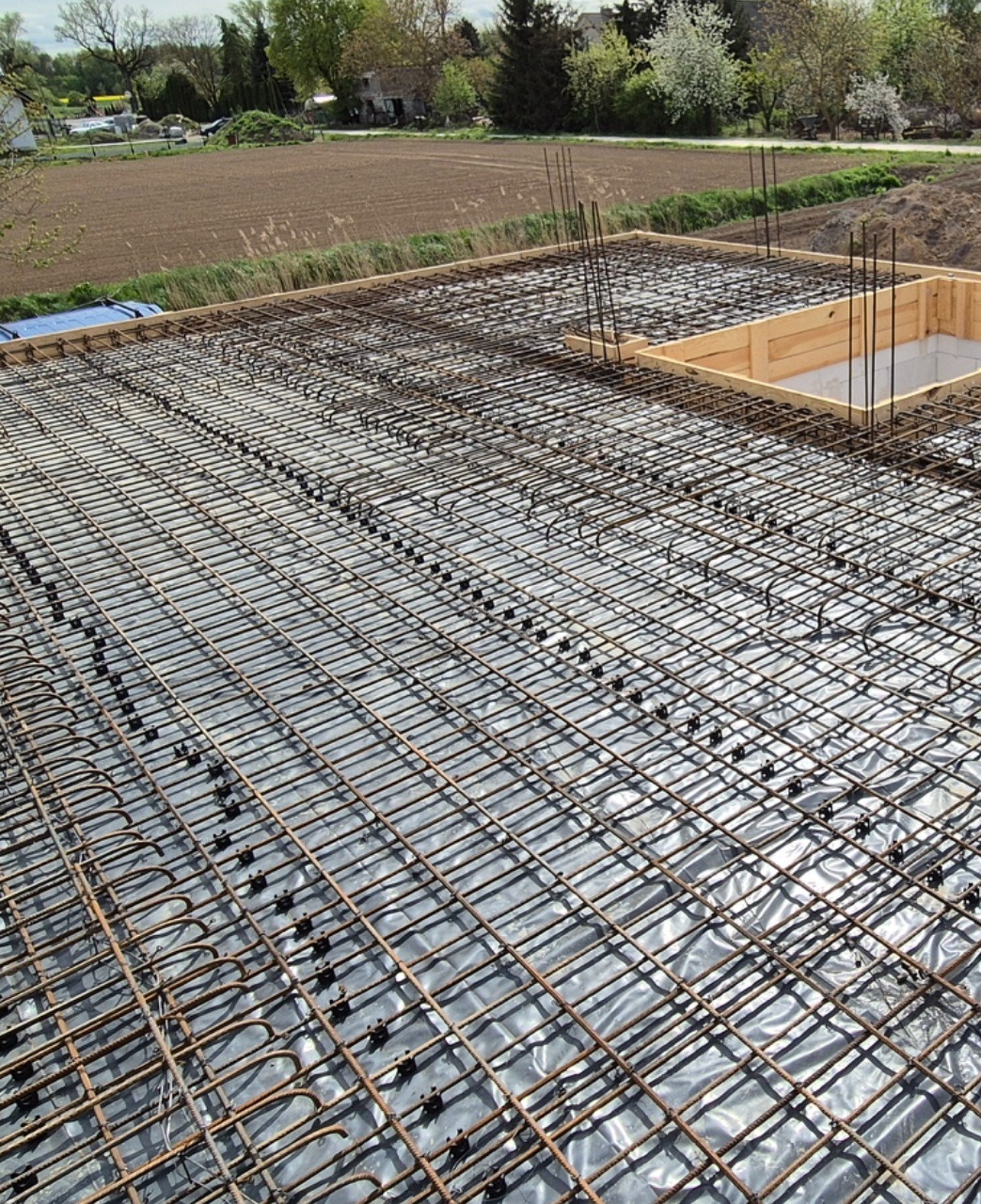 Siatka zbrojeniowa na folii, przygotowana do zalania betonem. Widoczne drewniane szalunki. Tło stanowią pola i drzewa.