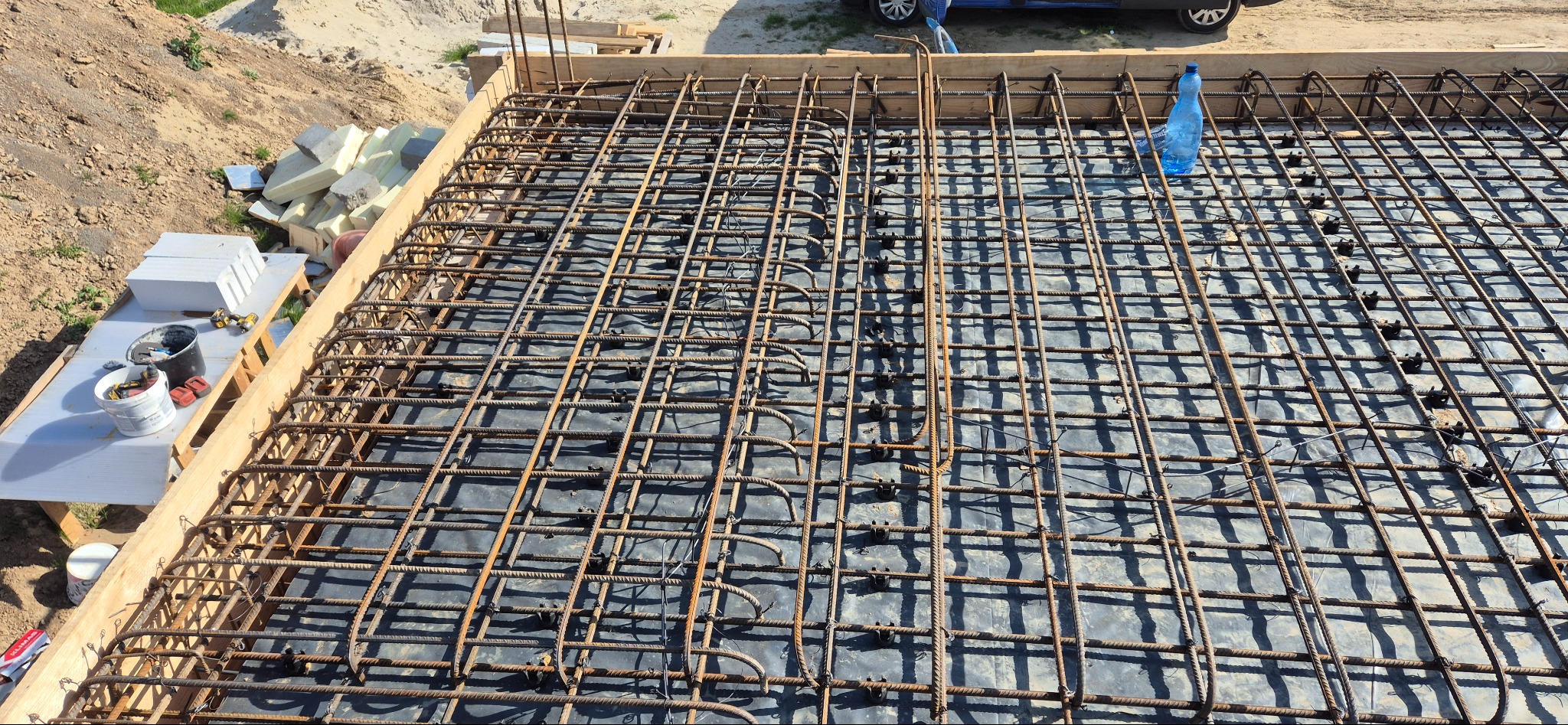Siatka zbrojeniowa ułożona na czarnej folii, przygotowana do zalania betonem. Widoczne drewniane szalunki i materiały budowlane w tle. Ujęcie z góry.