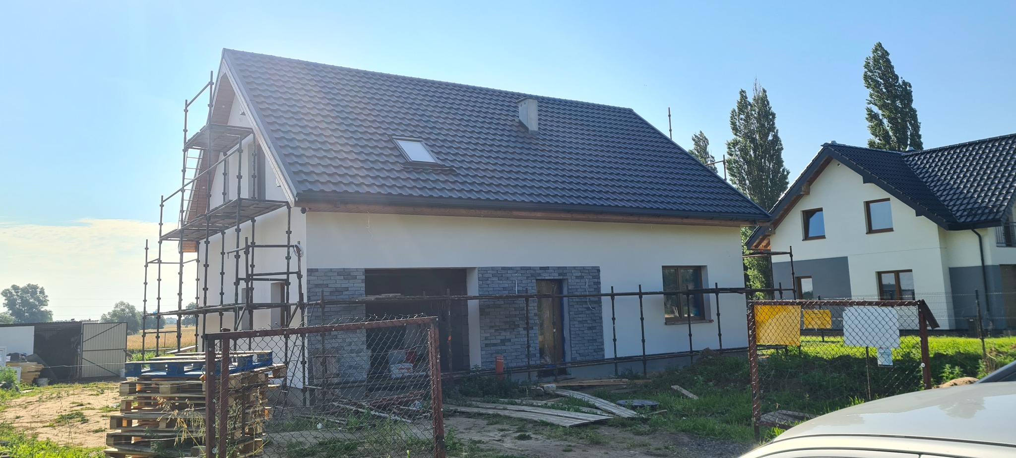 Dom jednorodzinny w trakcie wykończenia elewacji, z widocznym rusztowaniem i fragmentem drugiego budynku w tle. Dach pokryty ciemną dachówką, część elewacji wykończona cegłą.