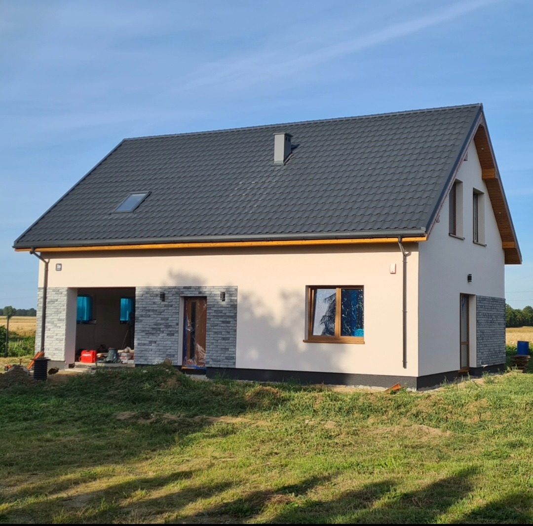 Nowoczesny dom jednorodzinny z szarą dachówką i elementami dekoracyjnymi z kamienia elewacyjnego. Widoczne okna z drewnianą stolarką i świeżo położona elewacja.