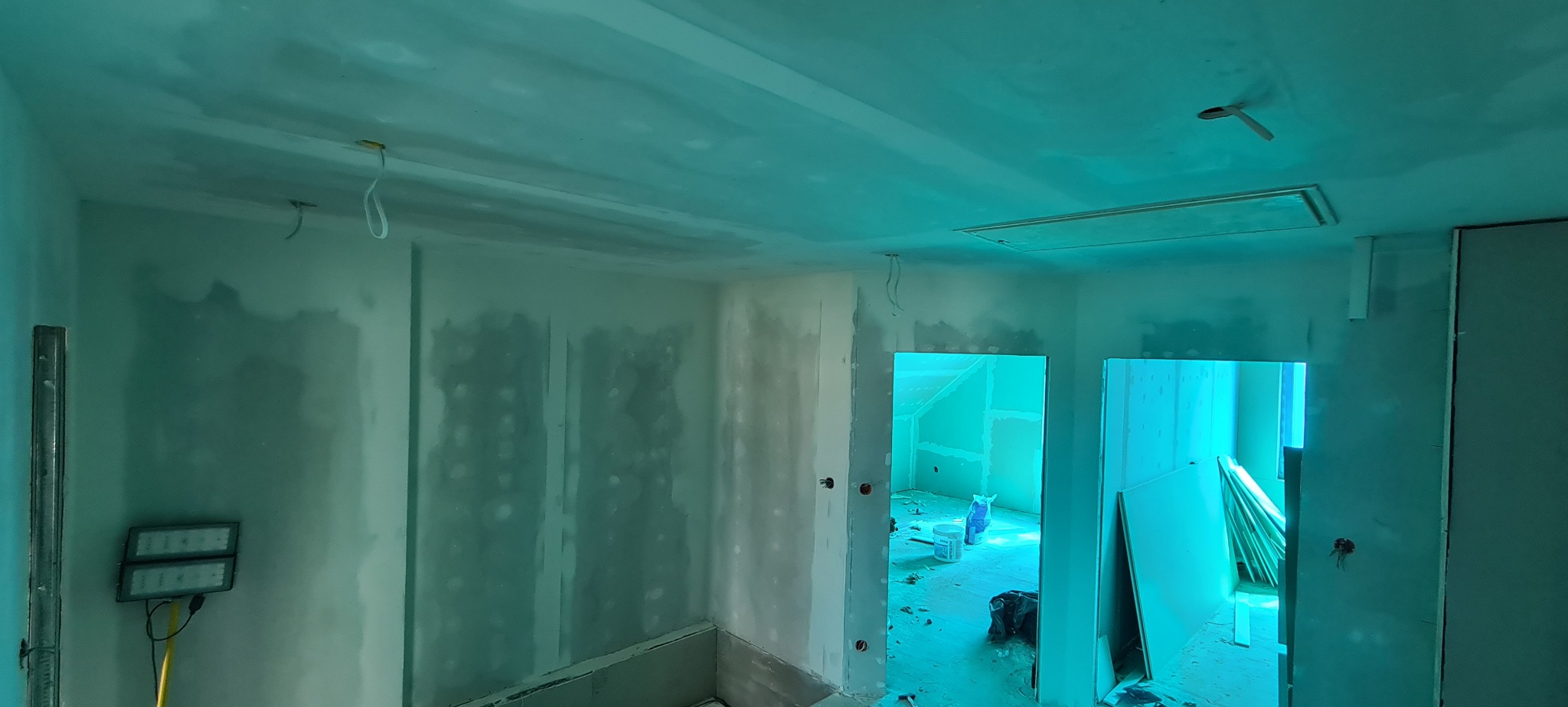 Wnętrze w trakcie remontu: ściany i sufit pokryte gładzią szpachlową, widoczne kable elektryczne, oświetlenie robocze. Prace wykończeniowe w nowym domu.