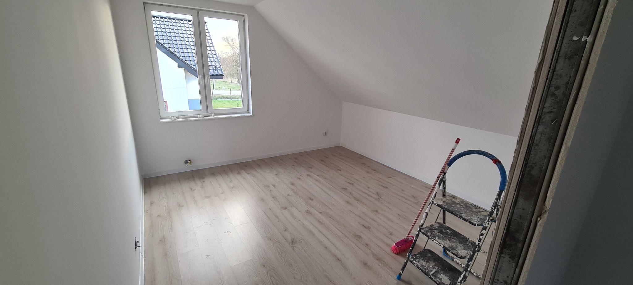 Poddasze po malowaniu ścian na biało, widoczna drabina malarska i okno z widokiem na dom z ciemnym dachem. Jasna podłoga z paneli.