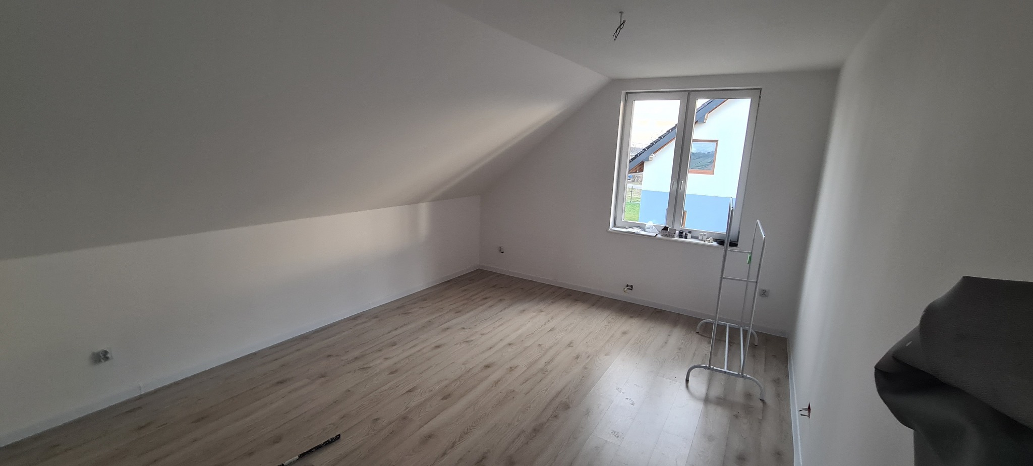 Jasne, świeżo pomalowane poddasze z oknem, ukazujące gładkie, białe ściany i podłogę z paneli. Widoczny stojak malarski. Minimalistyczny wystrój.