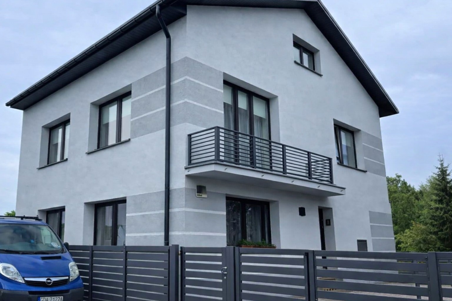 Dwukondygnacyjny dom z balkonem i szarą elewacją w pasy, widziany zza ogrodzenia. Na pierwszym planie zaparkowany niebieski samochód dostawczy.