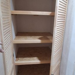 DamBud - Otwarta szafa wnękowa z drewnianymi lamelowymi drzwiami i półkami z płyty OSB. Widoczne wnętrze szafy i fragment podłogi z płytek.