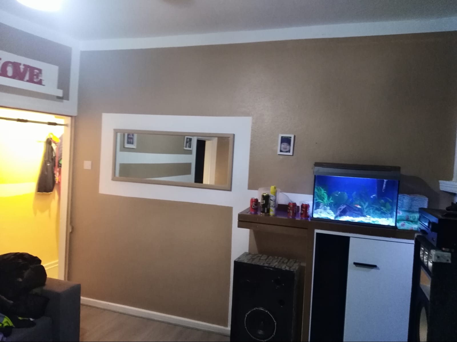 Beżowa ściana z lustrem w białej ramie i akwarium. Widoczny fragment pokoju z głośnikiem i szafką. Nad drzwiami napis LOVE.