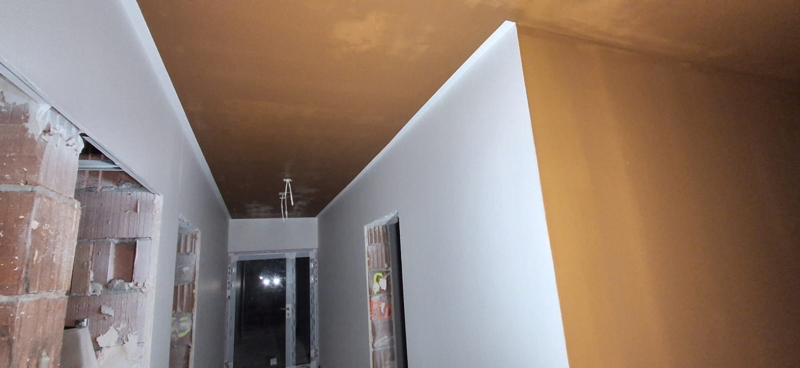 Wnętrze w trakcie prac wykończeniowych: gładkie, białe ściany kontrastują z surowym, brązowym sufitem. Widoczne fragmenty muru i instalacji elektrycznej.