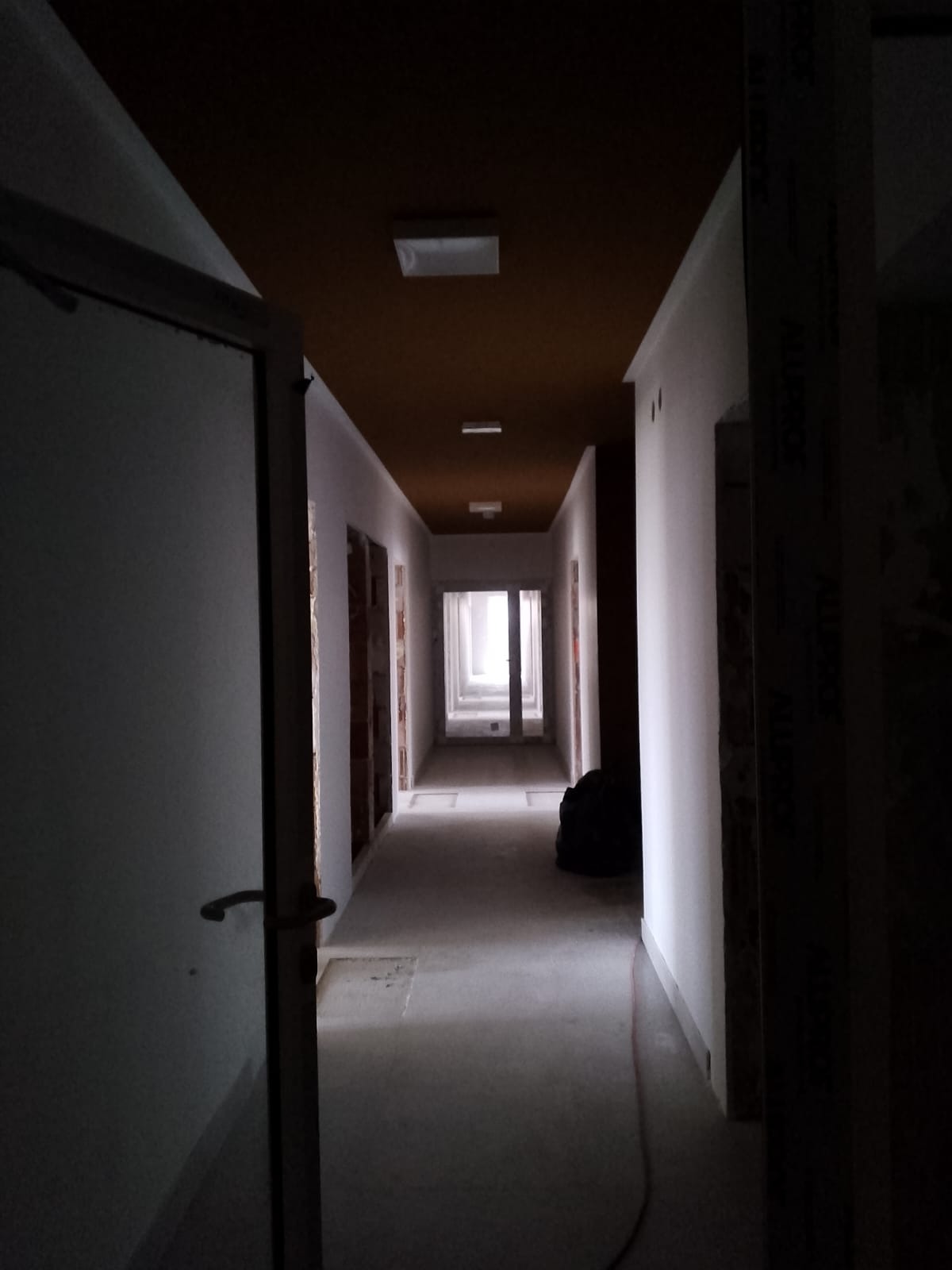 Długi korytarz w trakcie remontu. Widoczne surowe ściany, pomalowany sufit na brązowo i otwarte drzwi na końcu korytarza. Słabe oświetlenie.