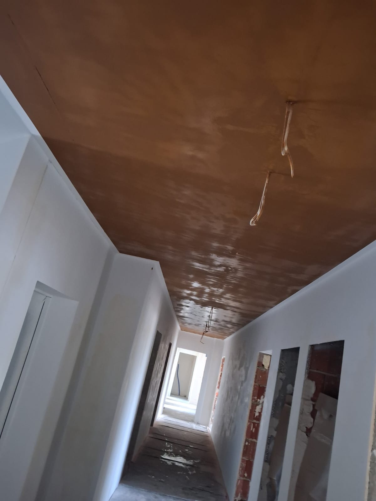 Korytarz w trakcie prac wykończeniowych: świeżo otynkowany sufit w odcieniach brązu, białe ściany, widoczne przewody. Ujęcie z dołu, perspektywa zbieżna.