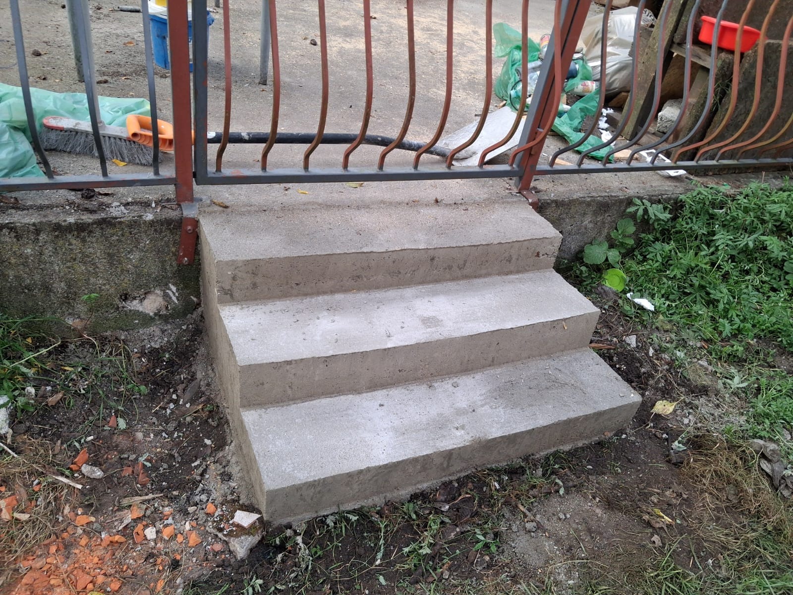 Nowe, betonowe schody wejściowe z trzema stopniami, prowadzące do metalowego ogrodzenia. Widoczne ślady świeżego betonu i otoczenie z ziemią i roślinnością.
