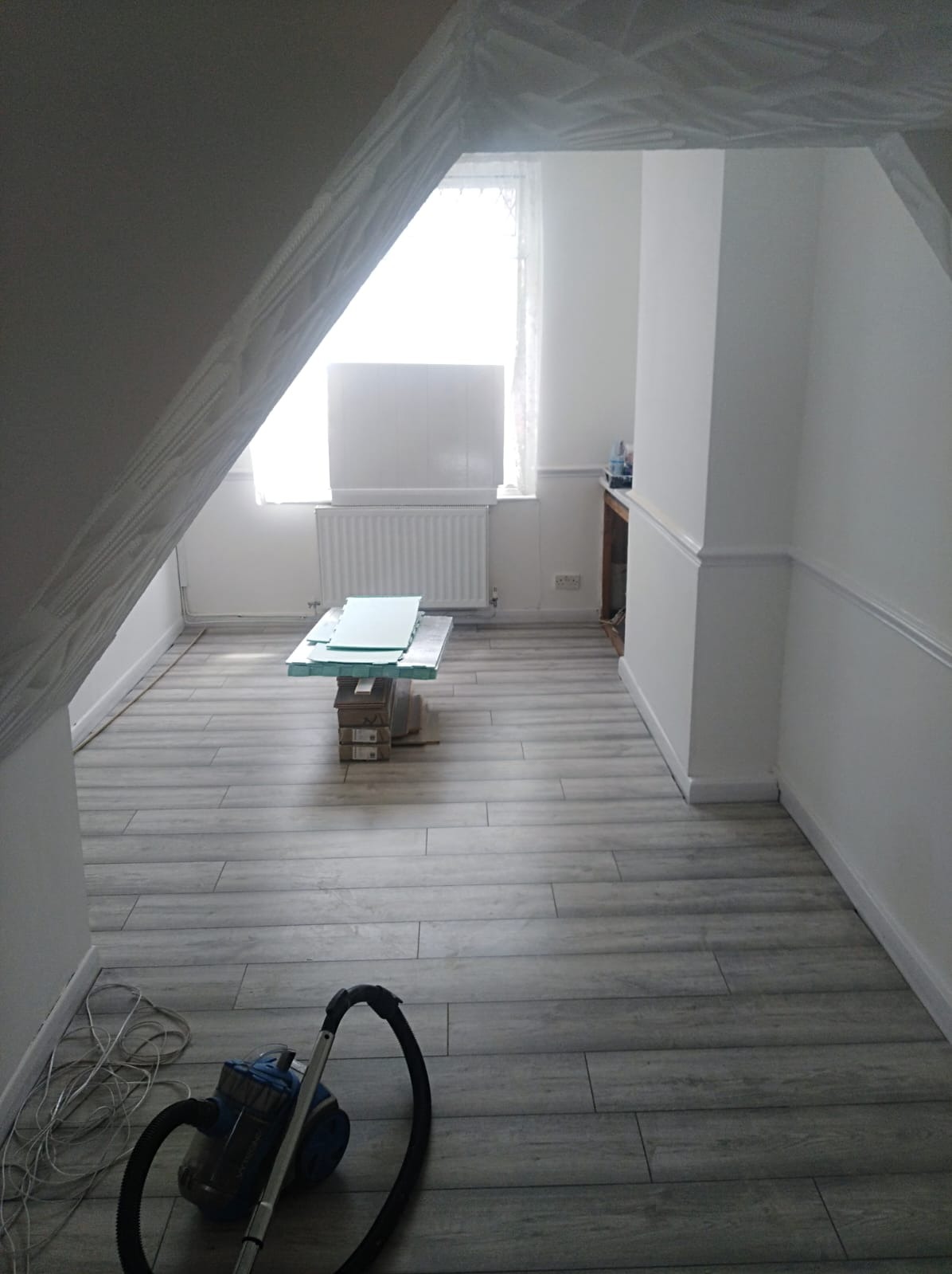 Pokój na poddaszu z nową podłogą z paneli imitujących drewno. Przy ścianie odkurzacz, w tle okno i materiały budowlane. Widok na wykończone wnętrze.