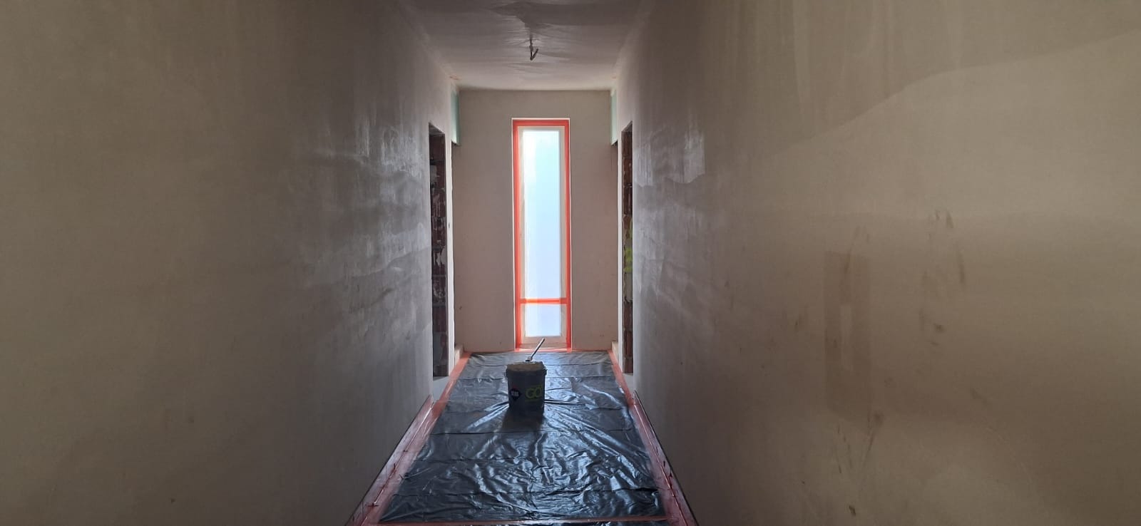 Korytarz w trakcie gładziowania ścian. Podłoga zabezpieczona folią, widoczne wiadro z zaprawą. Okno z pomarańczową ramą. Ściany szare, surowe.