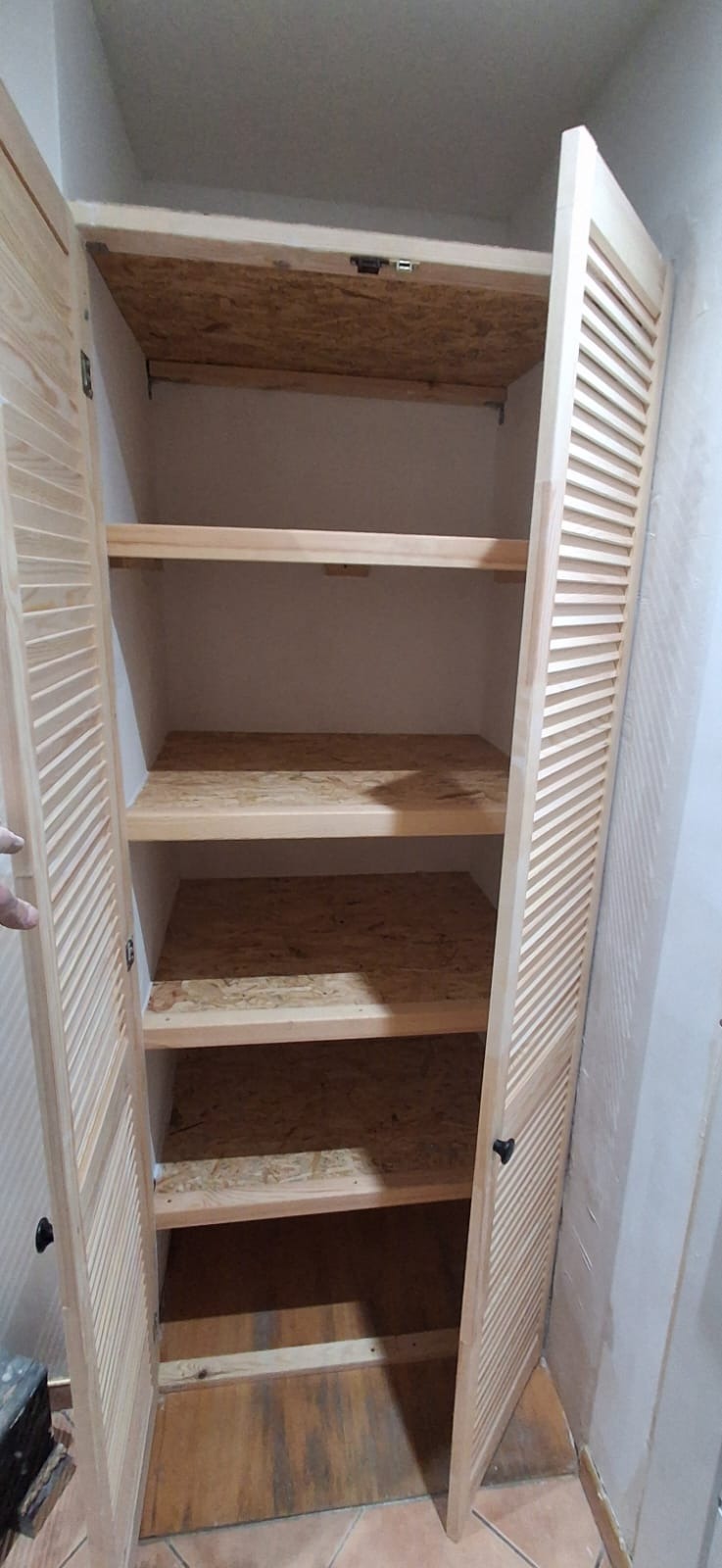 Otwarta szafa wnękowa z drewnianymi lamelowymi drzwiami i półkami z płyty OSB. Widoczne wnętrze szafy i fragment podłogi z płytek.