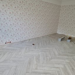 ML-System Marcin Langier - Ukończone układanie jasnych paneli podłogowych w jodełkę francuską w pokoju z tapetą w kwiaty. Widoczne narzędzia i puszki. Świeżo po remoncie.