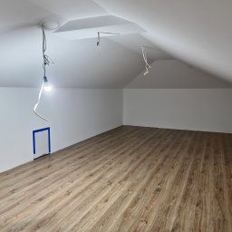 ML-System Marcin Langier - Poddasze po remoncie: świeżo ułożone panele podłogowe w odcieniach brązu, białe ściany i sufit ze skosami, widoczne kable i żarówka. Małe drzwiczki zabezpieczone taśmą.