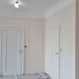 ML-System Marcin Langier - Białe, klasyczne wnętrze po remoncie: ściany z dekoracyjnymi listwami, dwie białe drzwi z złotymi klamkami, zabezpieczona podłoga. Minimalistyczny design.