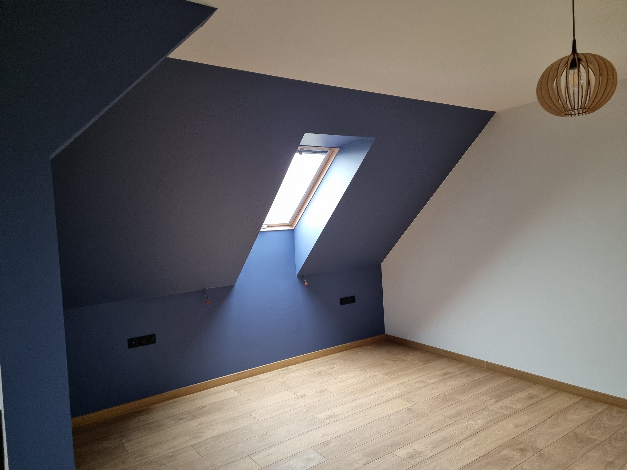 Pokój na poddaszu po remoncie: ściany pomalowane na granatowo i biało, drewniana podłoga, okno dachowe, nowoczesna lampa. Widoczne gniazdka elektryczne.