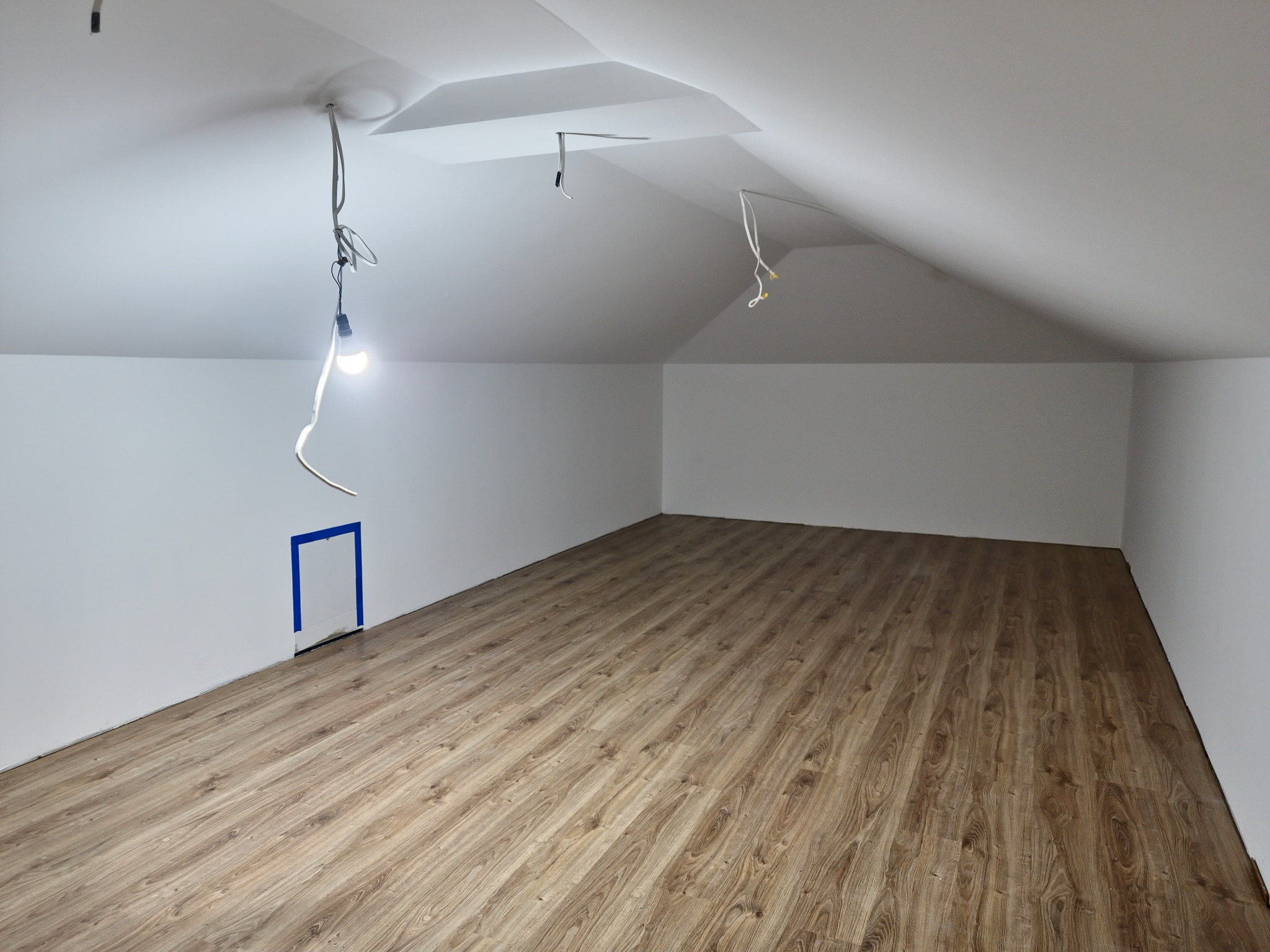 Poddasze po remoncie: świeżo ułożone panele podłogowe w odcieniach brązu, białe ściany i sufit ze skosami, widoczne kable i żarówka. Małe drzwiczki zabezpieczone taśmą.