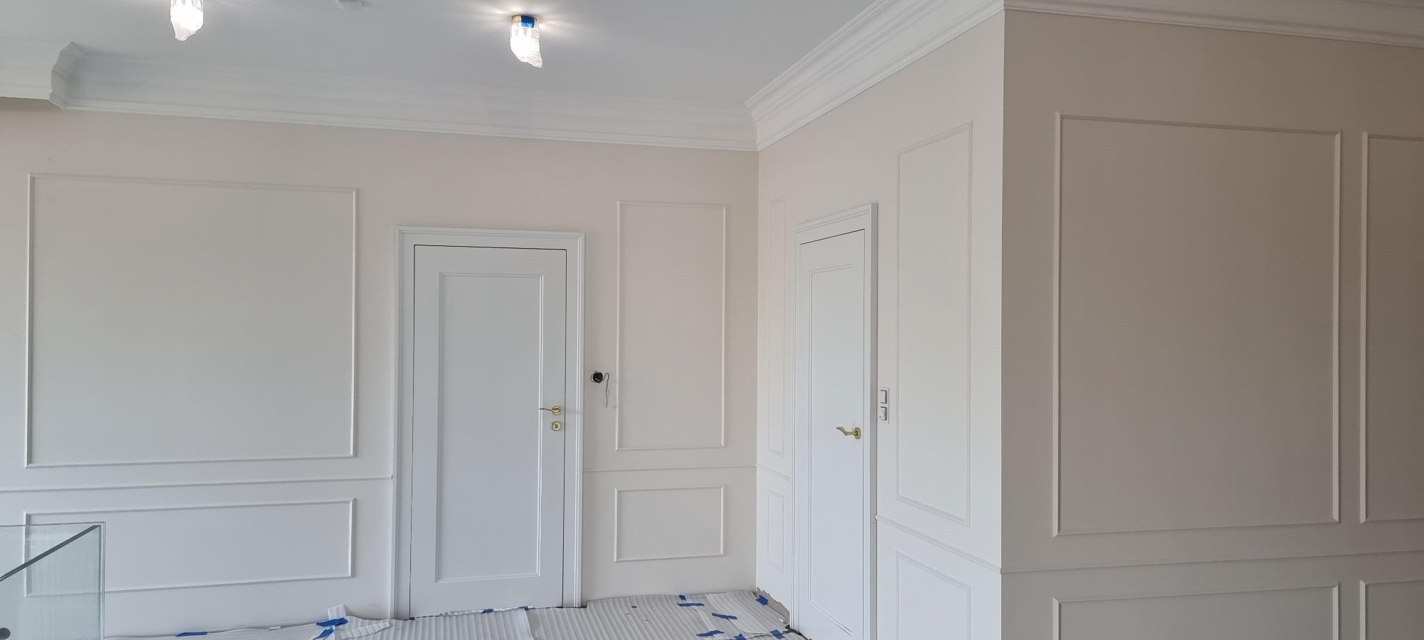 Białe, klasyczne wnętrze po remoncie: ściany z dekoracyjnymi listwami, dwie białe drzwi z złotymi klamkami, zabezpieczona podłoga. Minimalistyczny design.