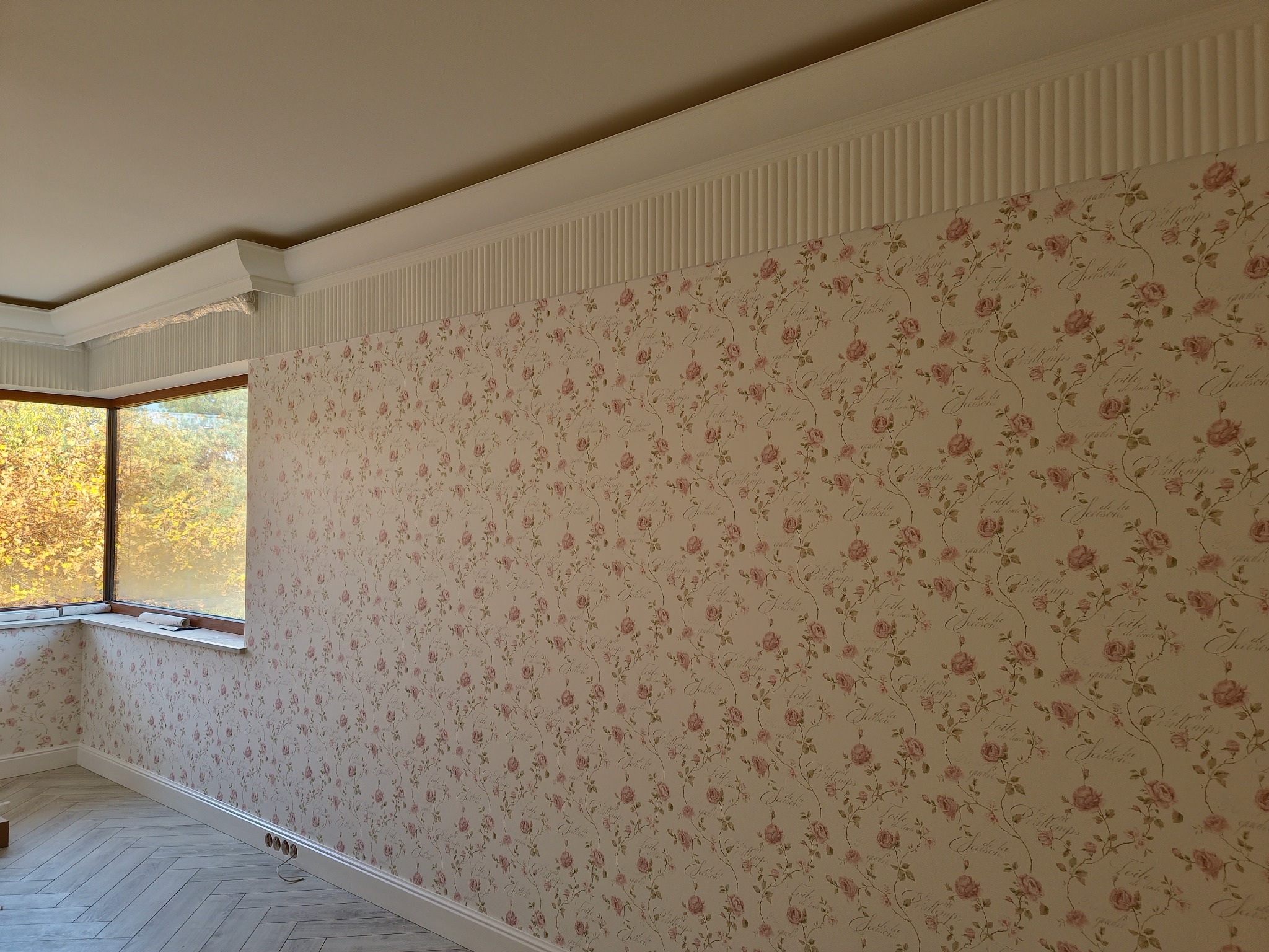 Pokój po remoncie: ściany w delikatne, różane tapety, białe listwy przypodłogowe i sufitowe, parkiet w jodełkę. Widok na okno z jesiennym krajobrazem.