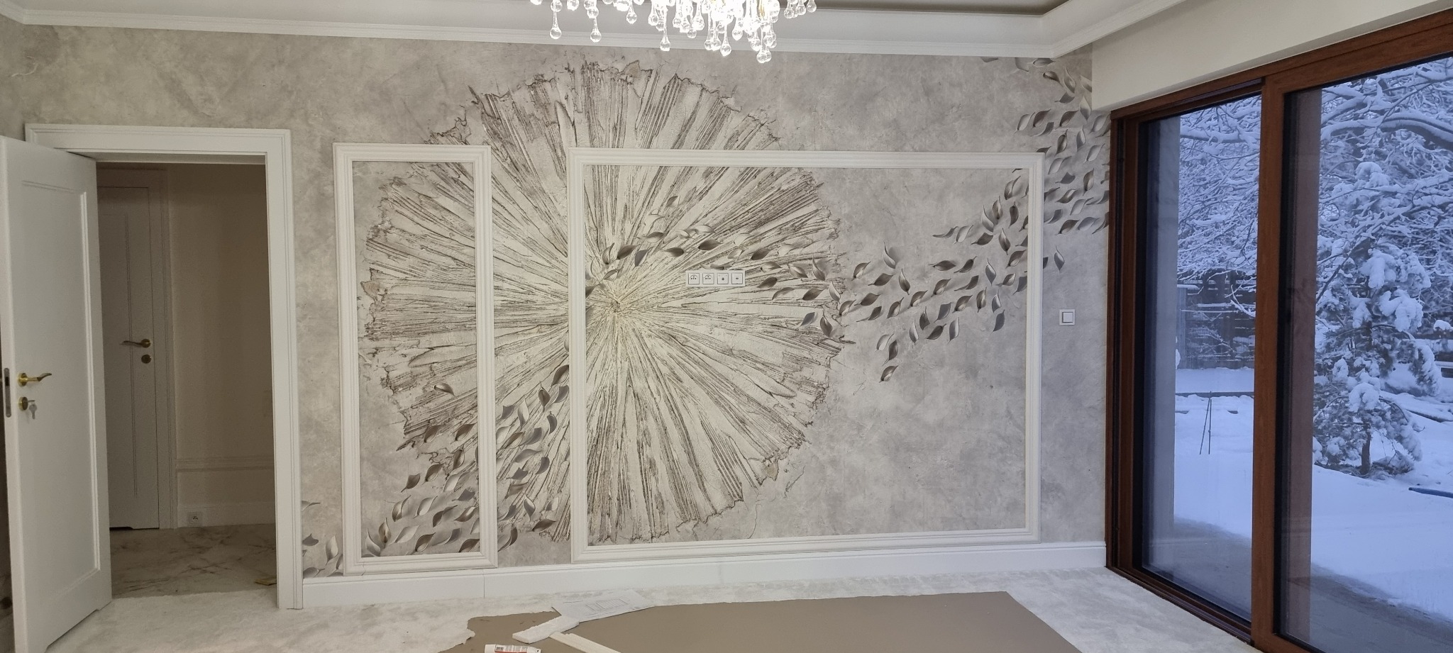 Eleganckie wykończenie ściany z dekoracyjną strukturą w kształcie rozety i motywem liści, obramowane listwami. Widok na zimowy krajobraz przez okno.