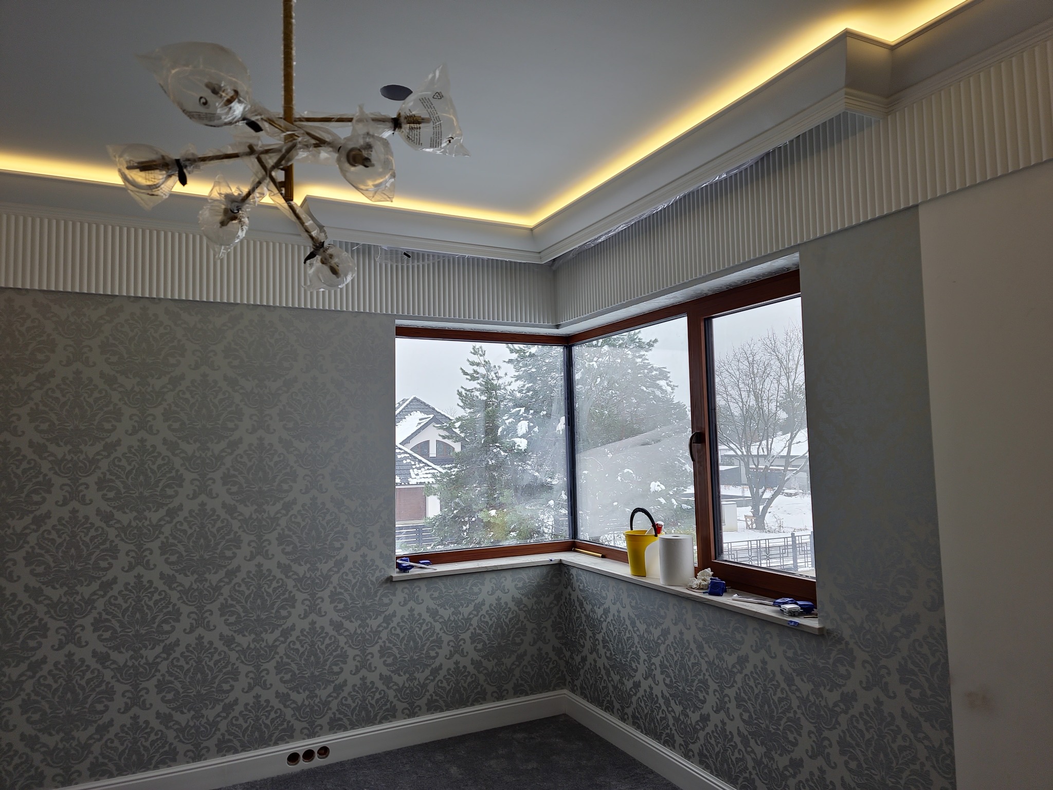 Pokój po remoncie: wzorzysta tapeta w odcieniach szarości, okno narożne z widokiem na zimowy krajobraz, sztukateria pod sufitem z oświetleniem LED. Lampa zabezpieczona folią.