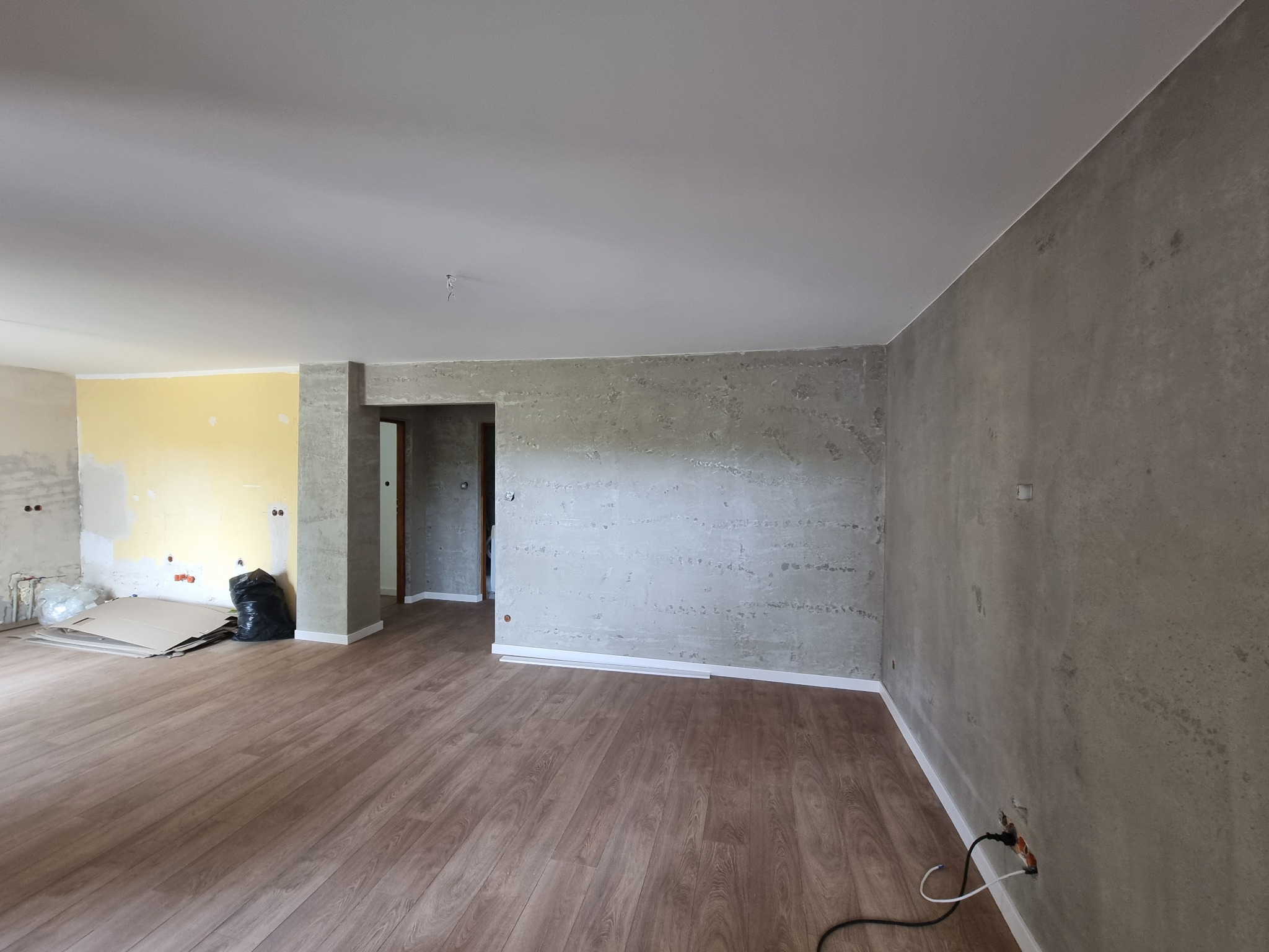 Wnętrze po remoncie: panele podłogowe, ściany z efektem betonu, fragment ściany przygotowanej do malowania. Widoczne ślady prac remontowych, kartony i worek na śmieci.