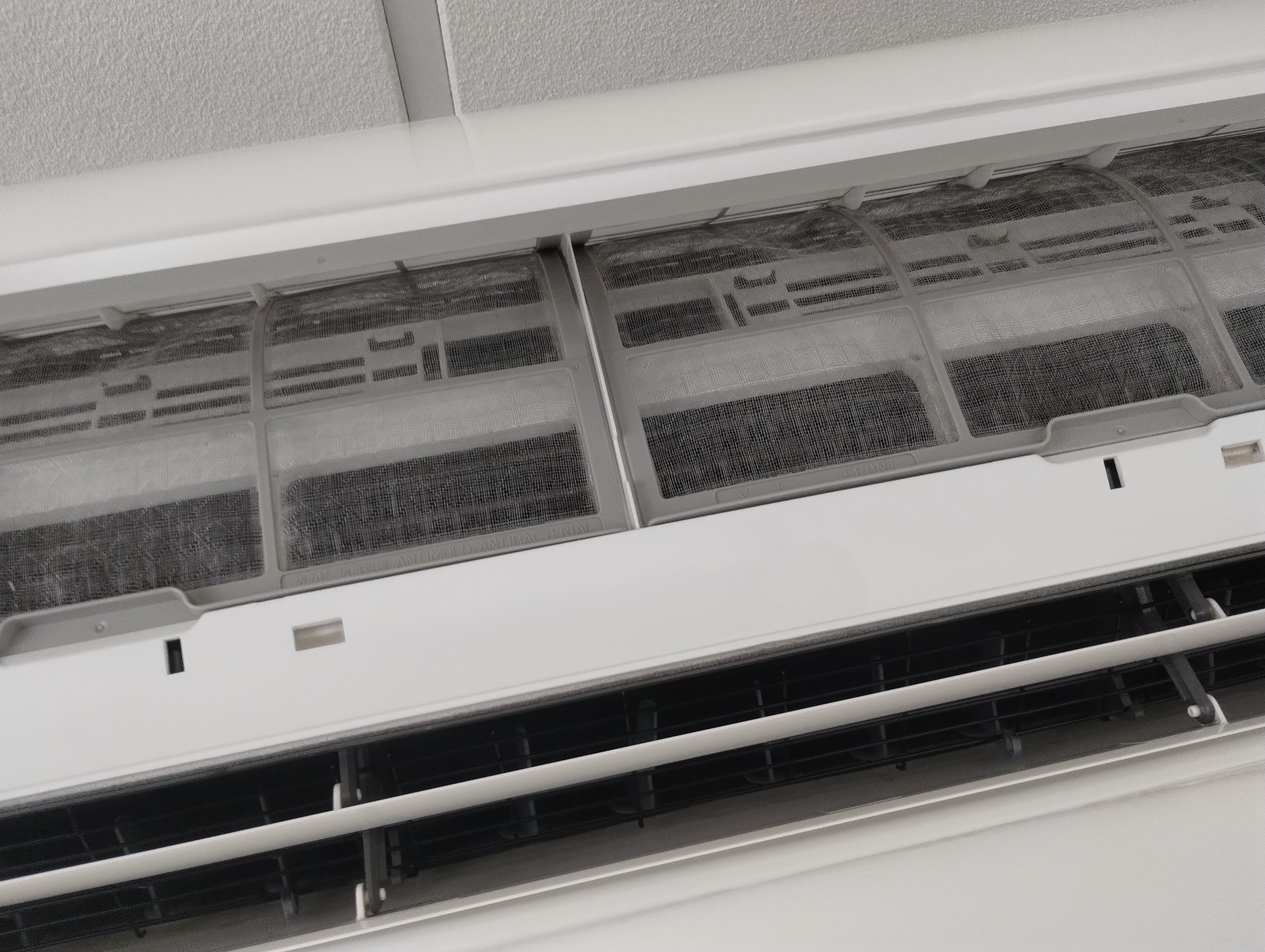 Biały klimatyzator ścienny z widocznymi filtrami, które wymagają czyszczenia. Widok z dołu na urządzenie zamontowane pod sufitem.