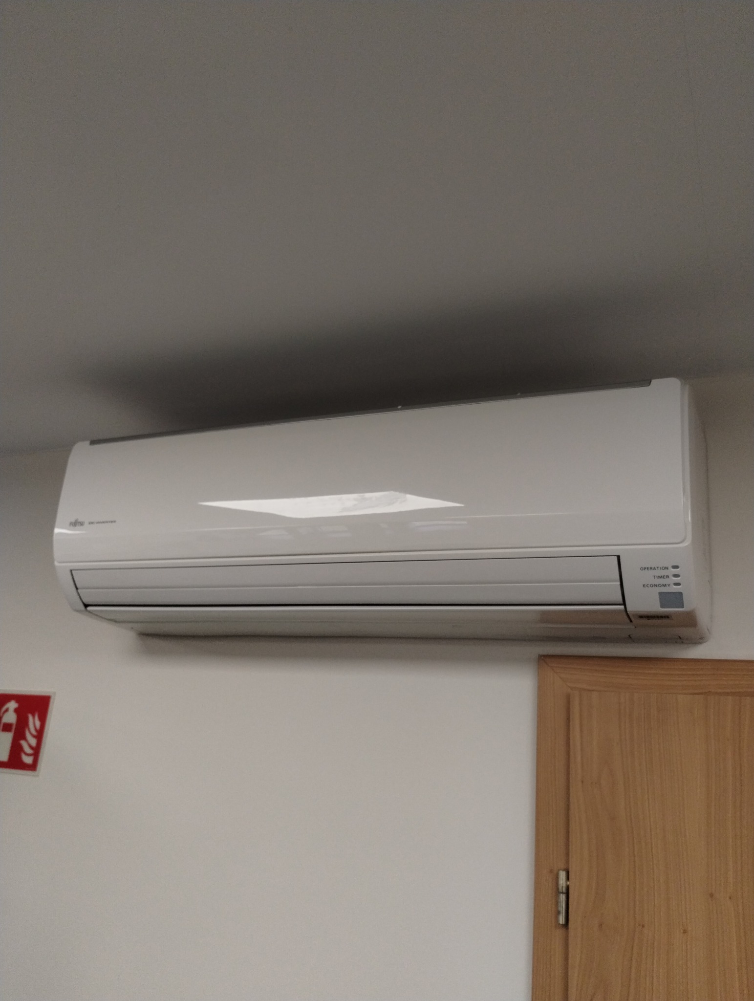 Biały klimatyzator Fujitsu zamontowany na ścianie, widoczne diody sygnalizacyjne, fragment drzwi z ościeżnicą i gaśnica na ścianie w tle. Ujęcie z dołu.
