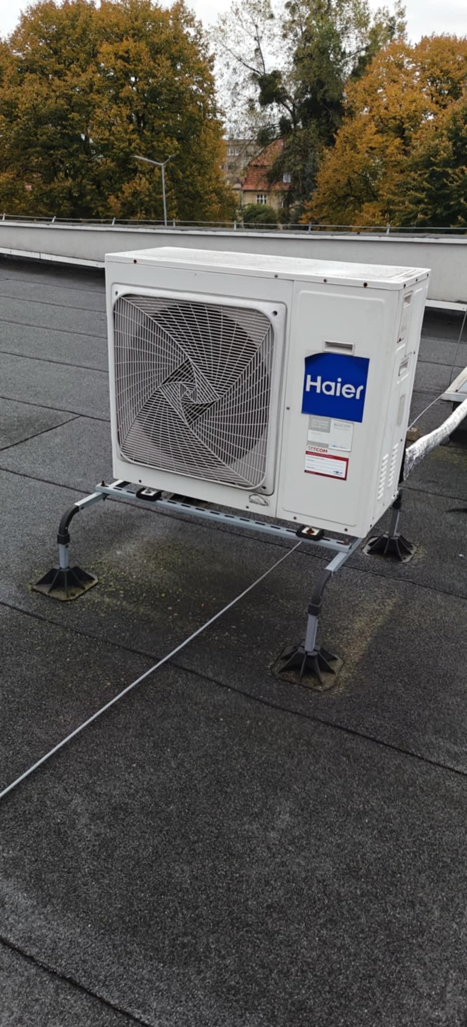 Biała jednostka zewnętrzna klimatyzacji Haier na dachu budynku, zamontowana na metalowej konstrukcji. W tle drzewa z jesiennymi liśćmi i budynki.