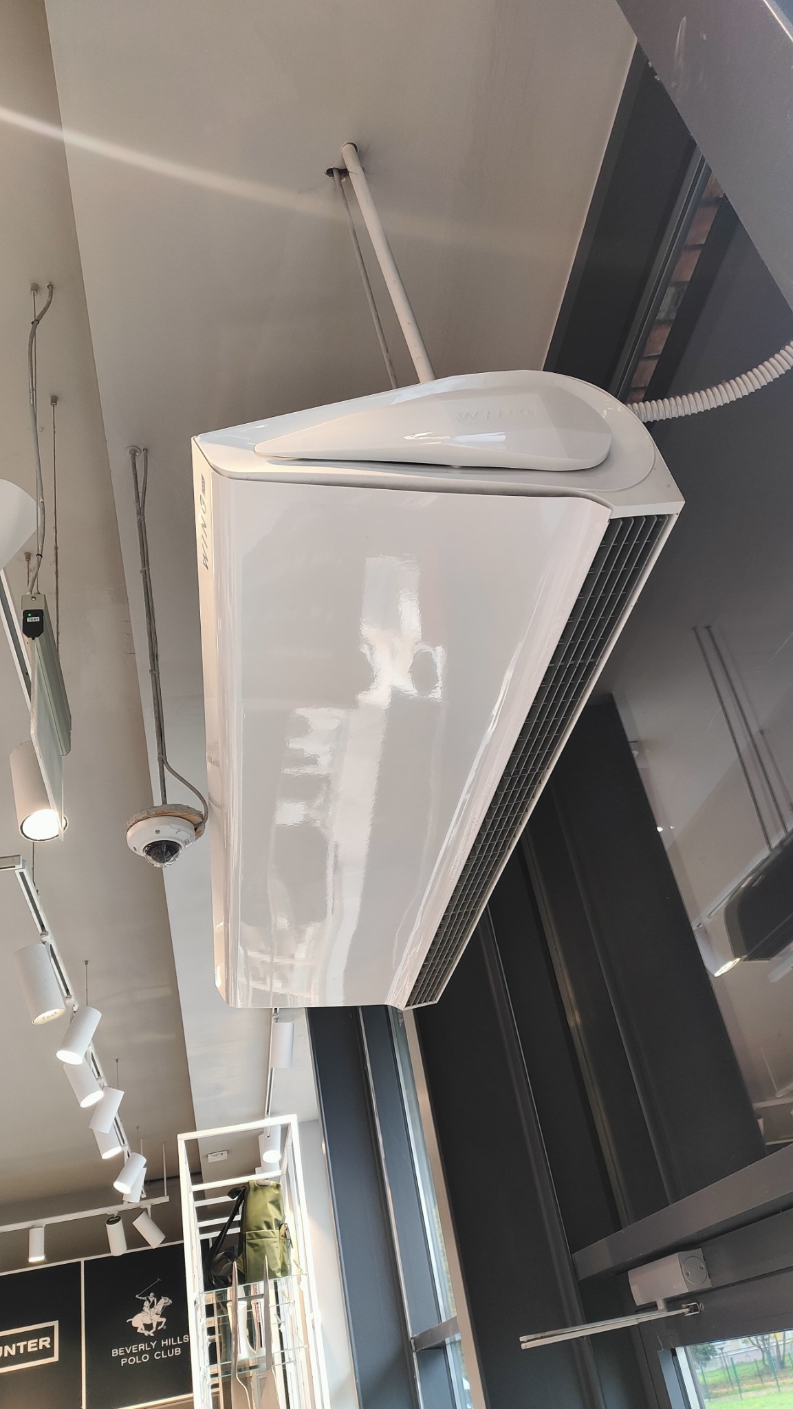 Biała klimatyzacja sufitowa w nowoczesnym wnętrzu sklepu odzieżowego, widoczna z dołu pod kątem. Widoczne oświetlenie i elementy wystroju.