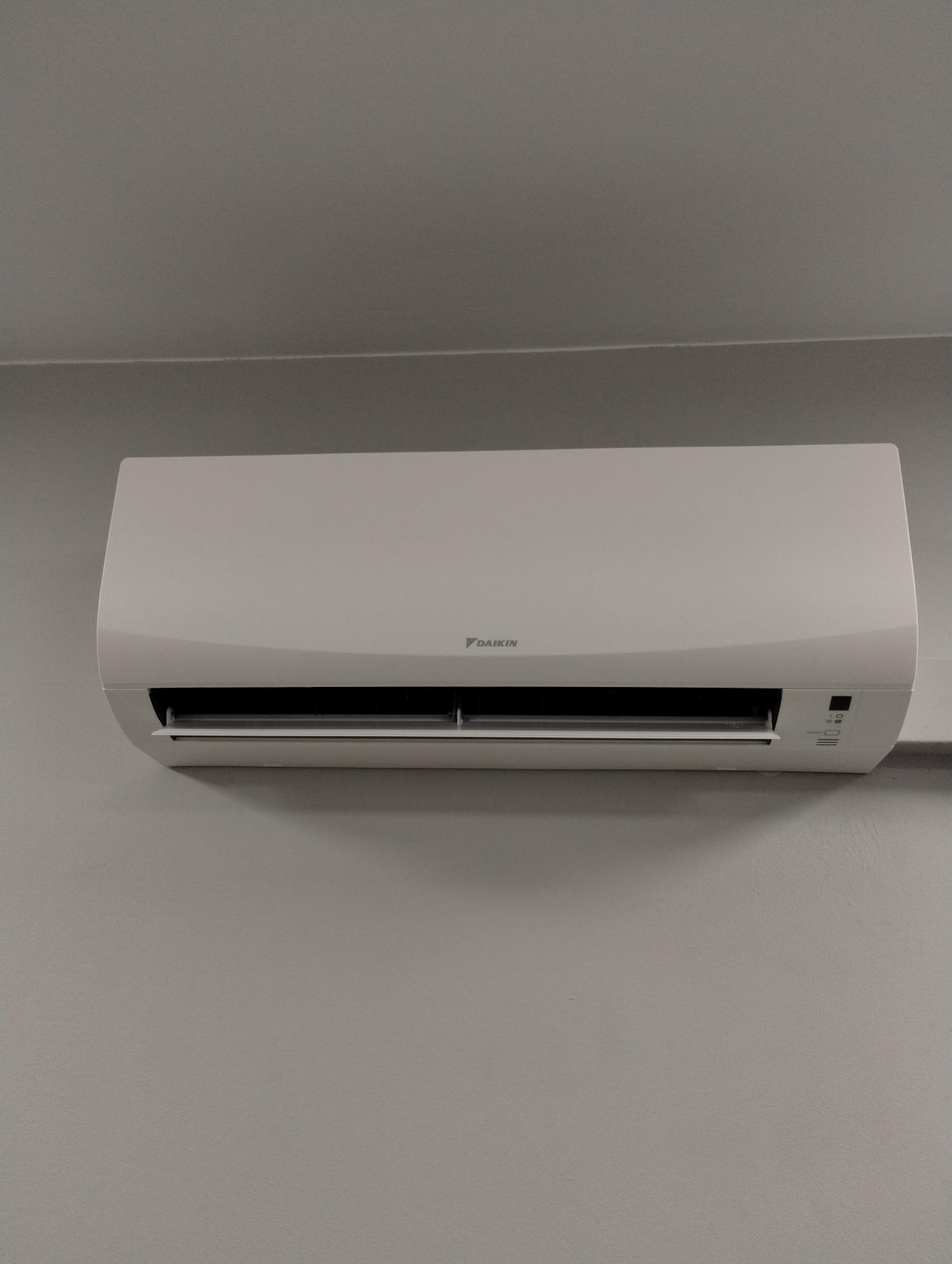 Biały klimatyzator Daikin zamontowany na szarej ścianie. Widoczne detale obudowy i panel sterowania. Minimalistyczny design, neutralne tło.