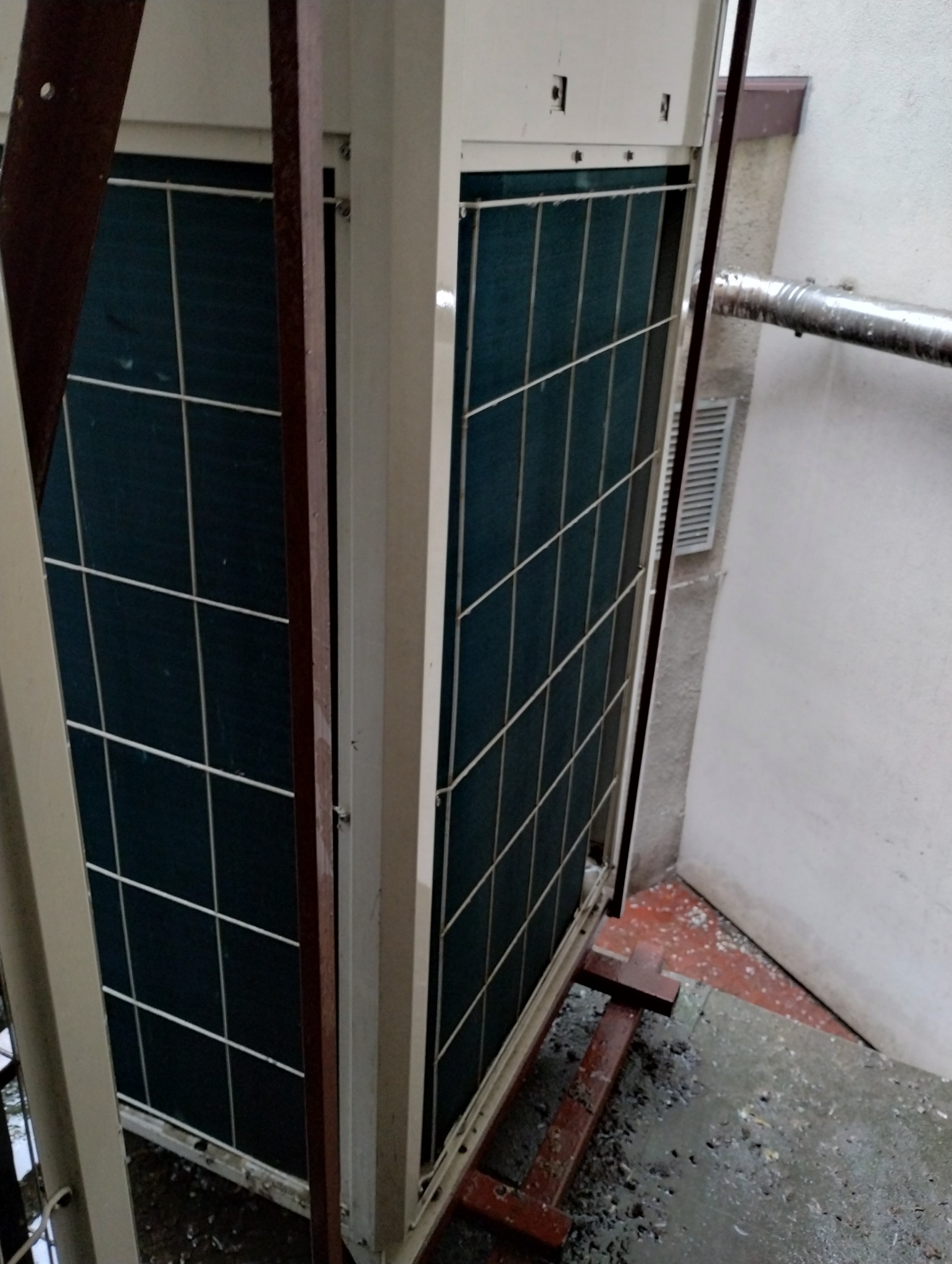 Zewnętrzna jednostka klimatyzacji, widok z boku, na metalowej konstrukcji. Widoczne kratki wentylacyjne i fragment ściany budynku.