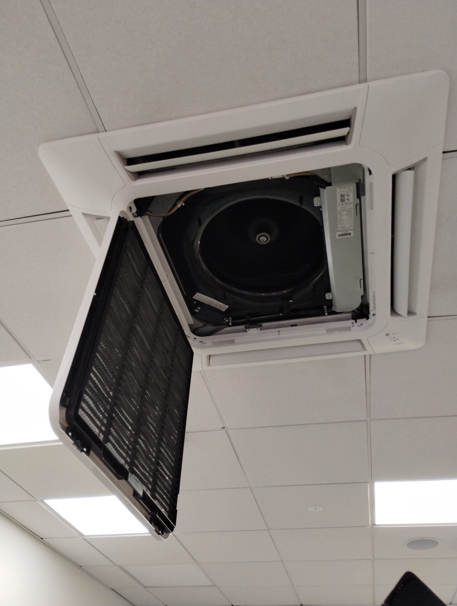 Kasetonowa klimatyzacja sufitowa z otwartym filtrem, widoczny wentylator. Biała obudowa na tle sufitu podwieszanego. Ujęcie z dołu.